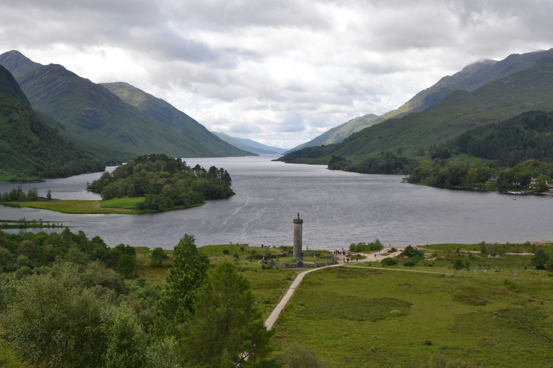 Loch Shiel, Scotland, with a monument on a grassy shore, an island, and mountains under a cloudy sky.