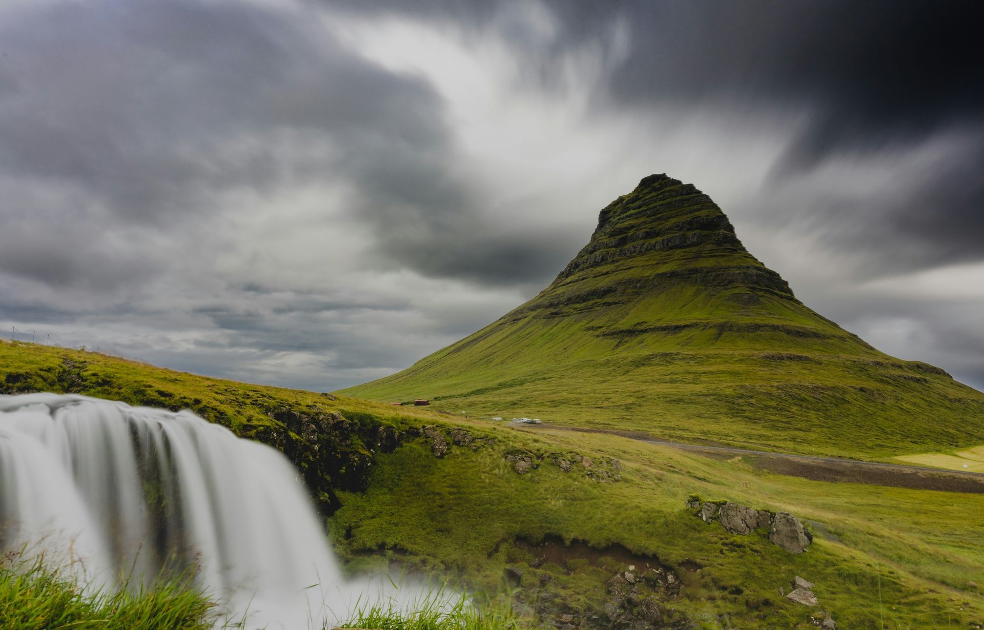 Waterfall cascading into a grassy field with a cone-shaped green mountain under a cloudy sky in Snæfellsnes, Iceland.
