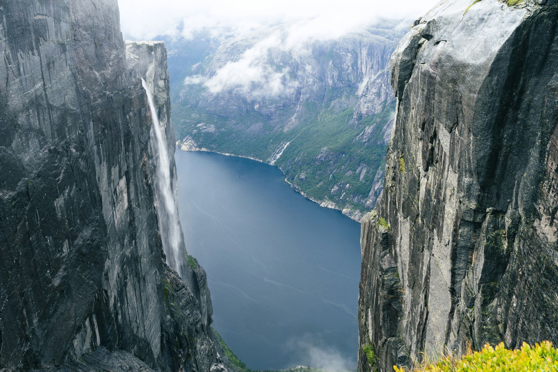 A waterfall cascades between tall cliffs into Lysefjord, with mountains and clouds in the distance in Norway.