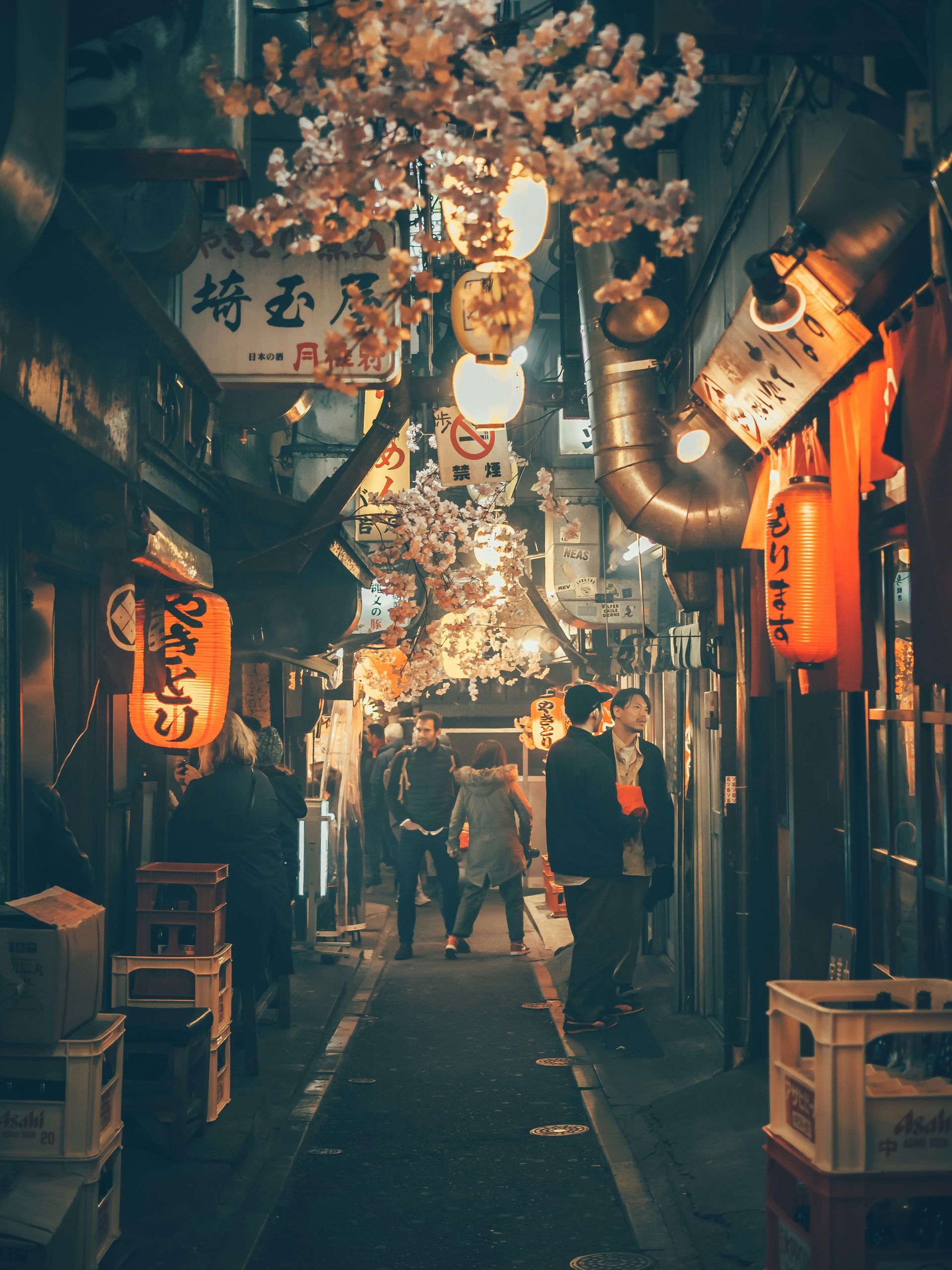 Narrow alley at night in Tokyo with lanterns, blossoming trees, and people walking.