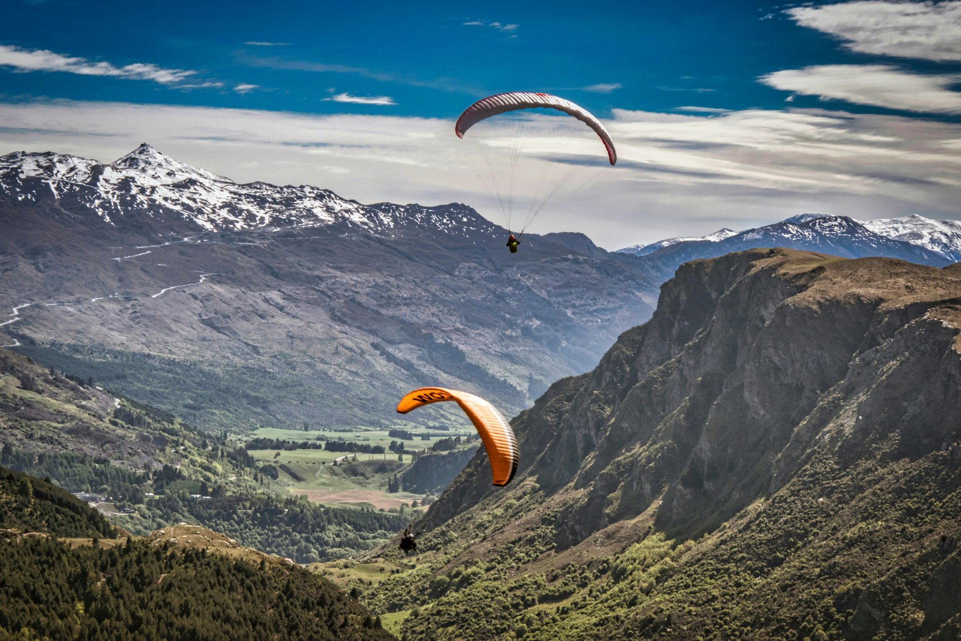 Two paragliders are flying over a mountain valley in Queenstown, New Zealand.