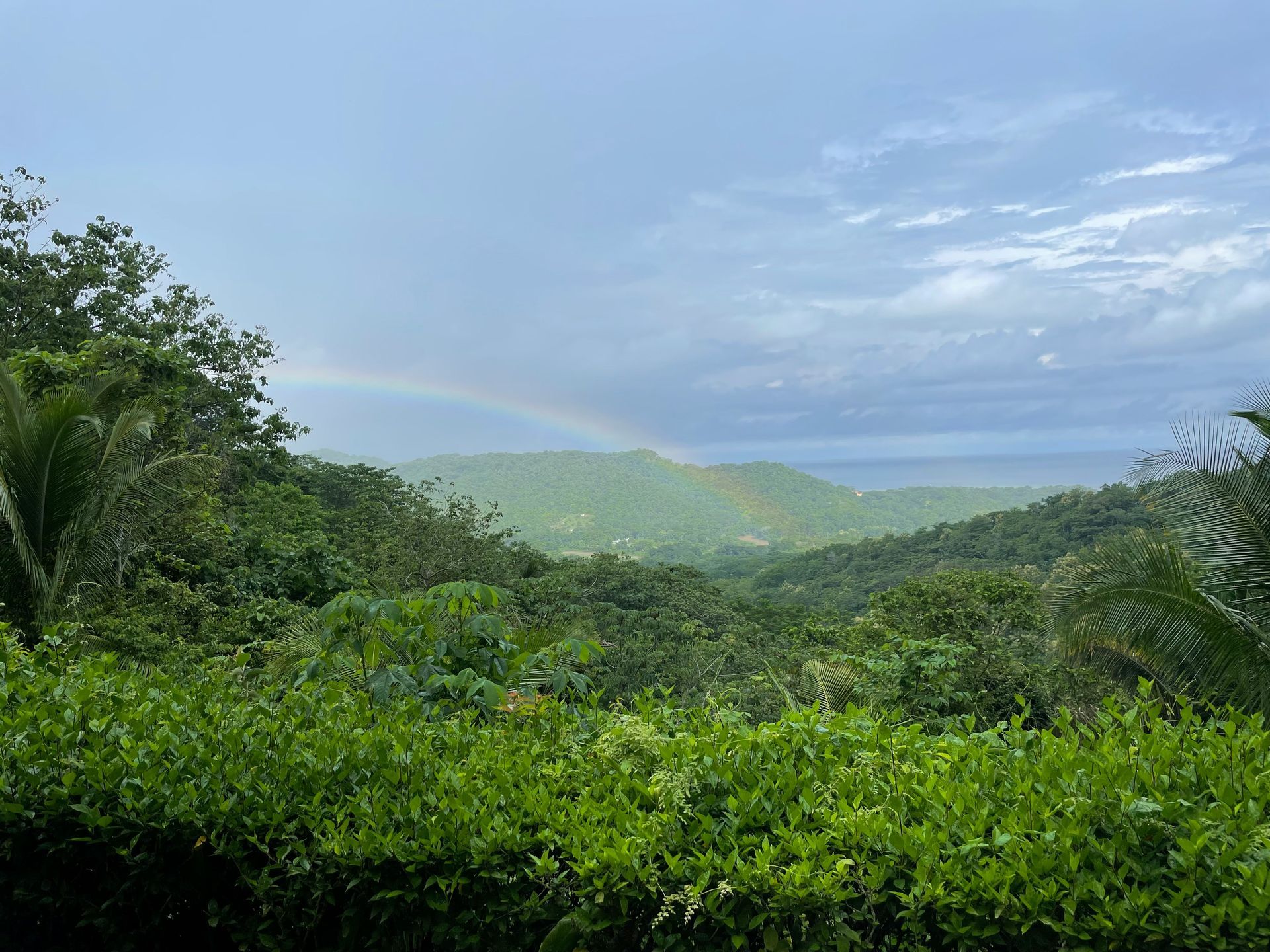 A vibrant rainbow arches over a lush, green valley, with a glimpse of the ocean visible under a cloudy sky in the Osa Peninsula, Costa Rica.