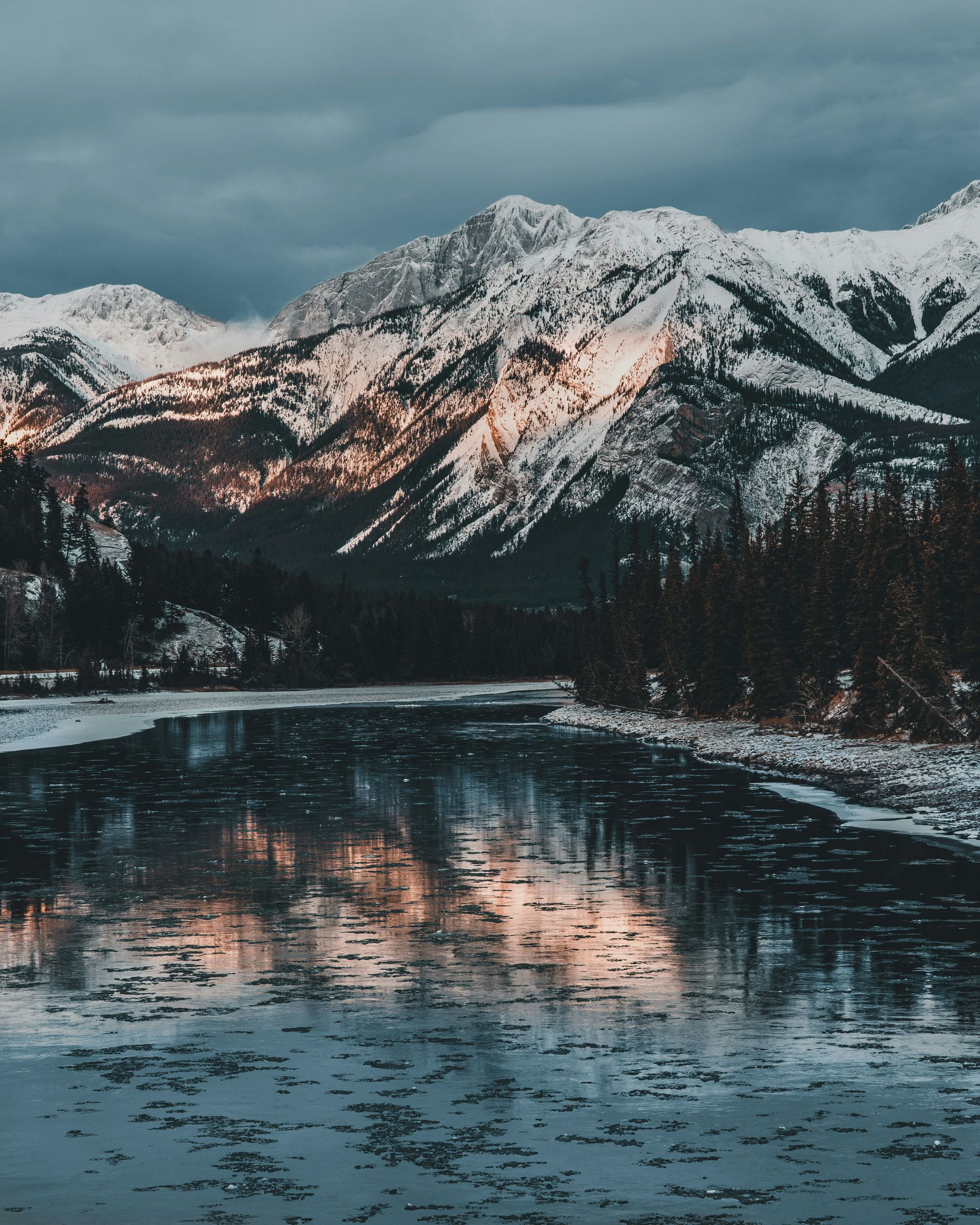 Snow-capped mountains reflecting in a partially frozen river, with sunlight hitting the peaks in Jasper, Canada.