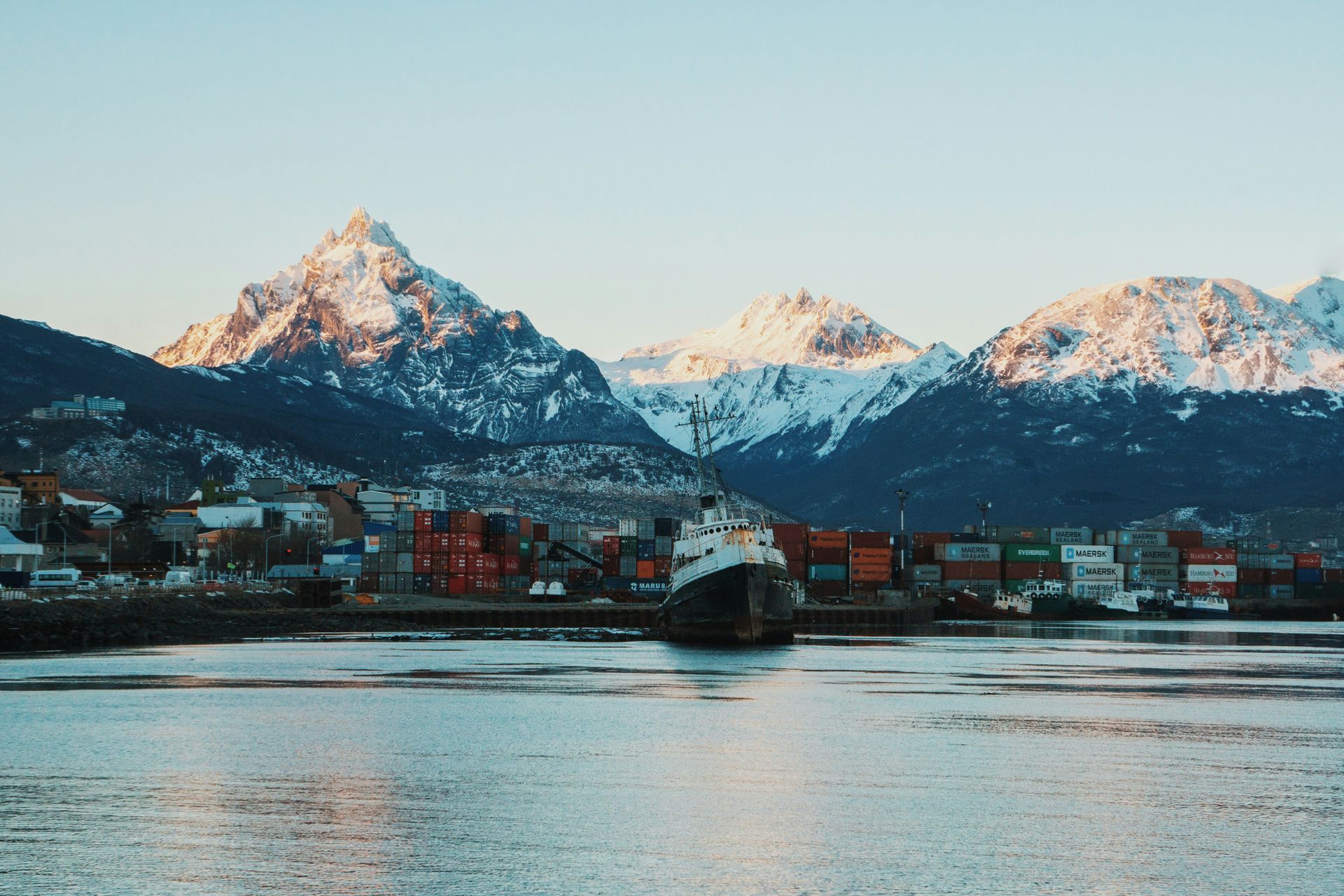 Snow-capped mountains frame a harbor scene; a cargo ship sails past colorful containers in Ushuaia, Argentina.