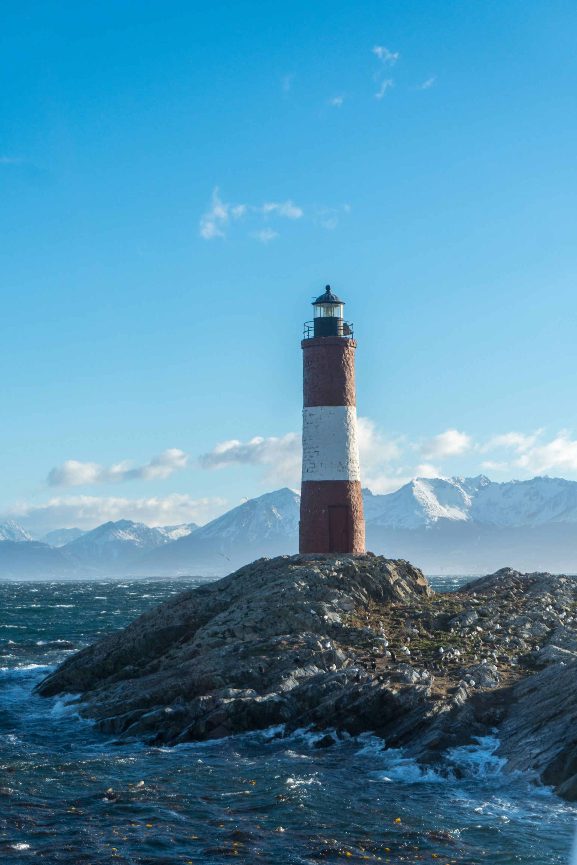 Red and white lighthouse on rocky outcrop, waves crashing, snow-capped mountains in the background in Ushuaia, Argentina.