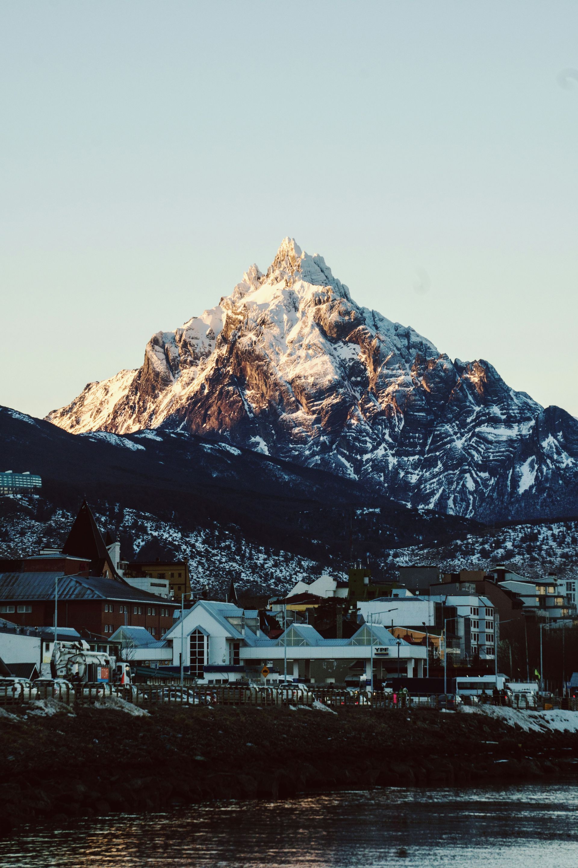 Snow-capped mountain towering over a waterfront town at sunrise, with buildings in Ushuaia, Argentina.
