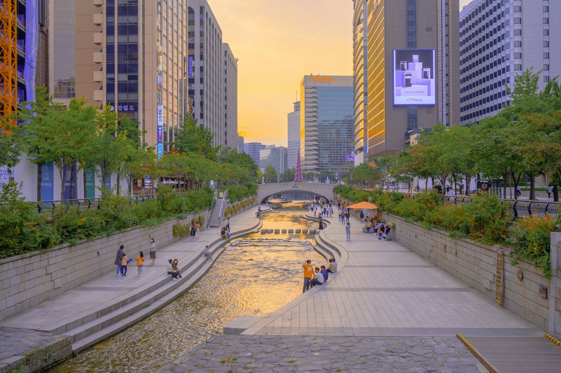 Stream flowing through Seoul, Korea, flanked by walking paths and buildings. Sunset colors reflect on the water.