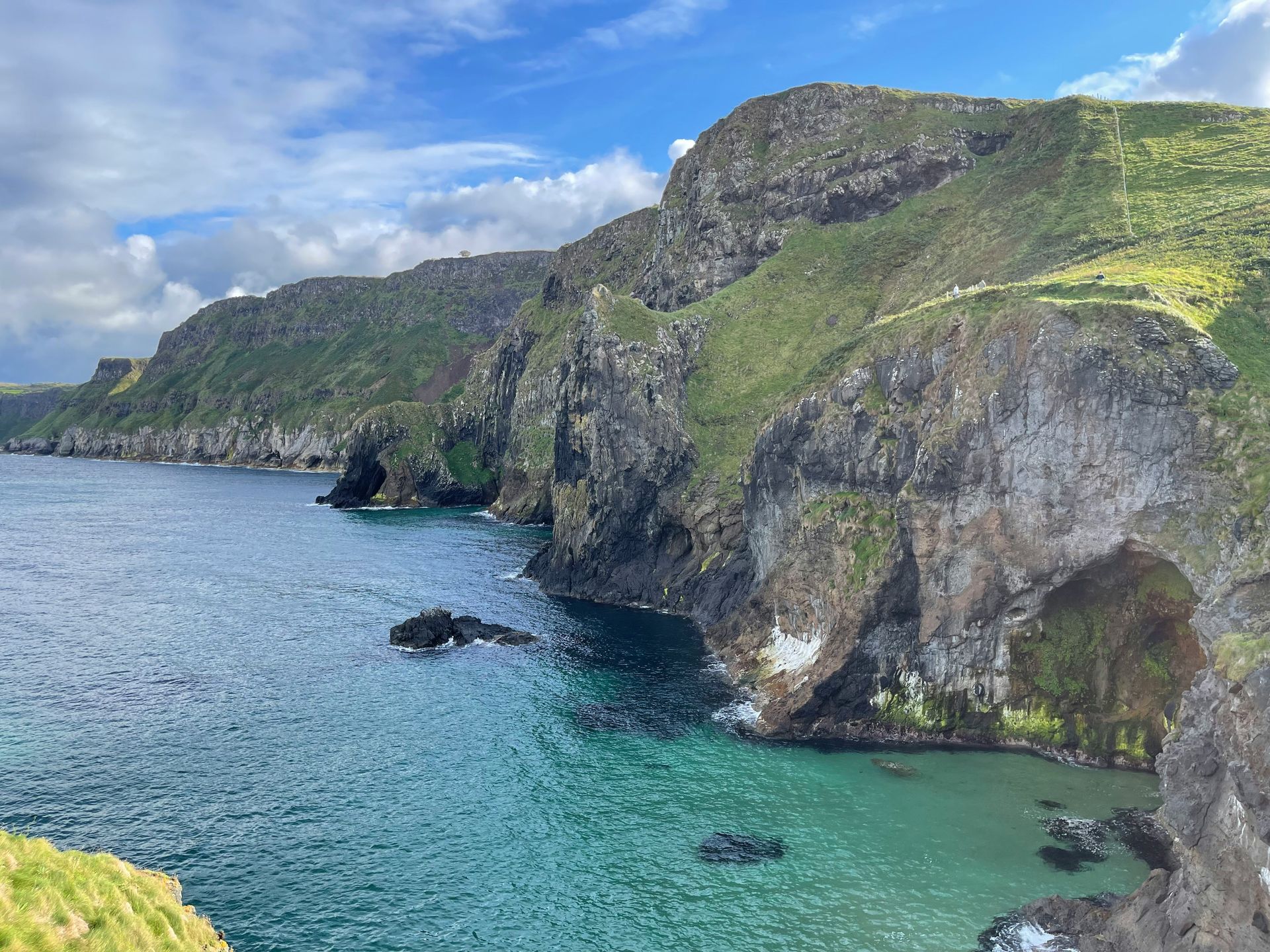 Rocky coastline with turquoise water, cliffs, and green hills under a cloudy sky at Carrick-a-Rede, Northern Ireland. 