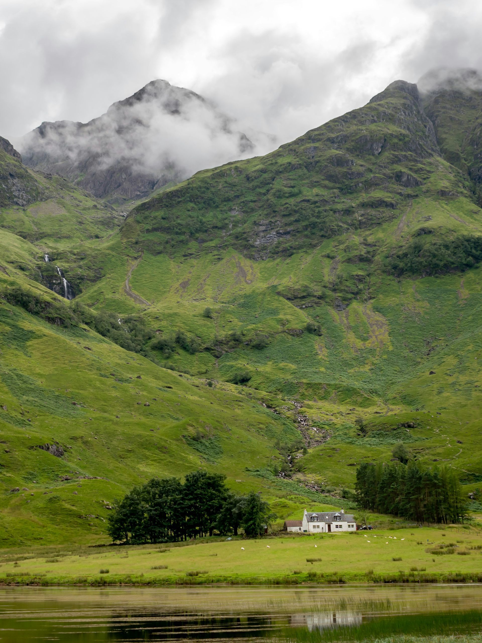 Rolling green hills and a small white house nestle at the base of a misty mountain range in Glencoe, Scotland.