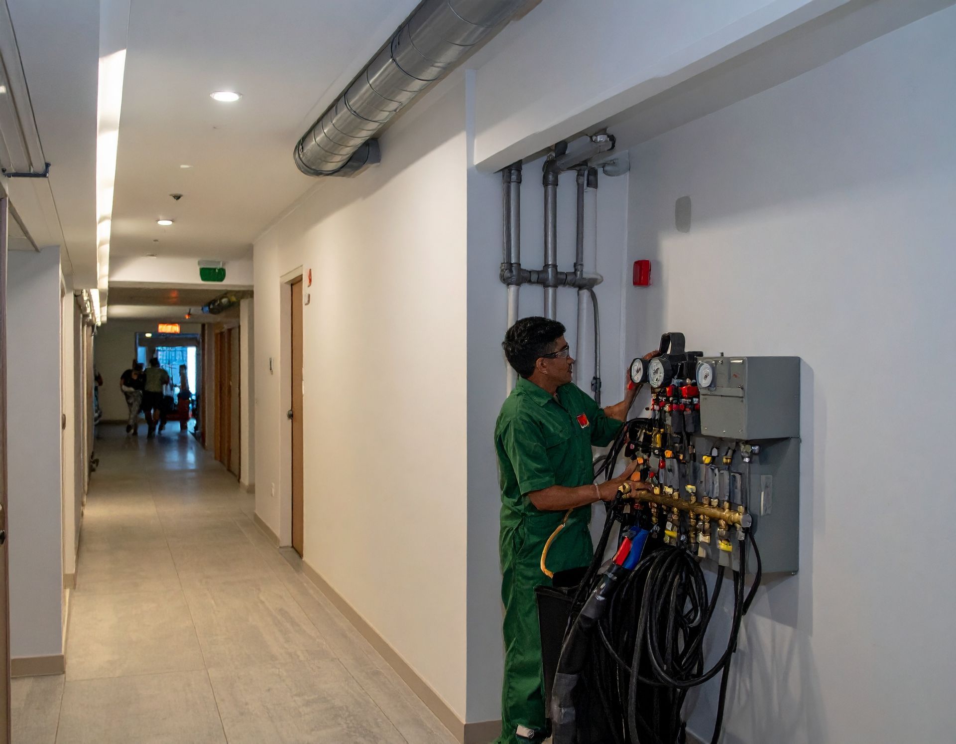 Worker in green uniform checking electrical panels in a bright hallway; people visible in the distance.