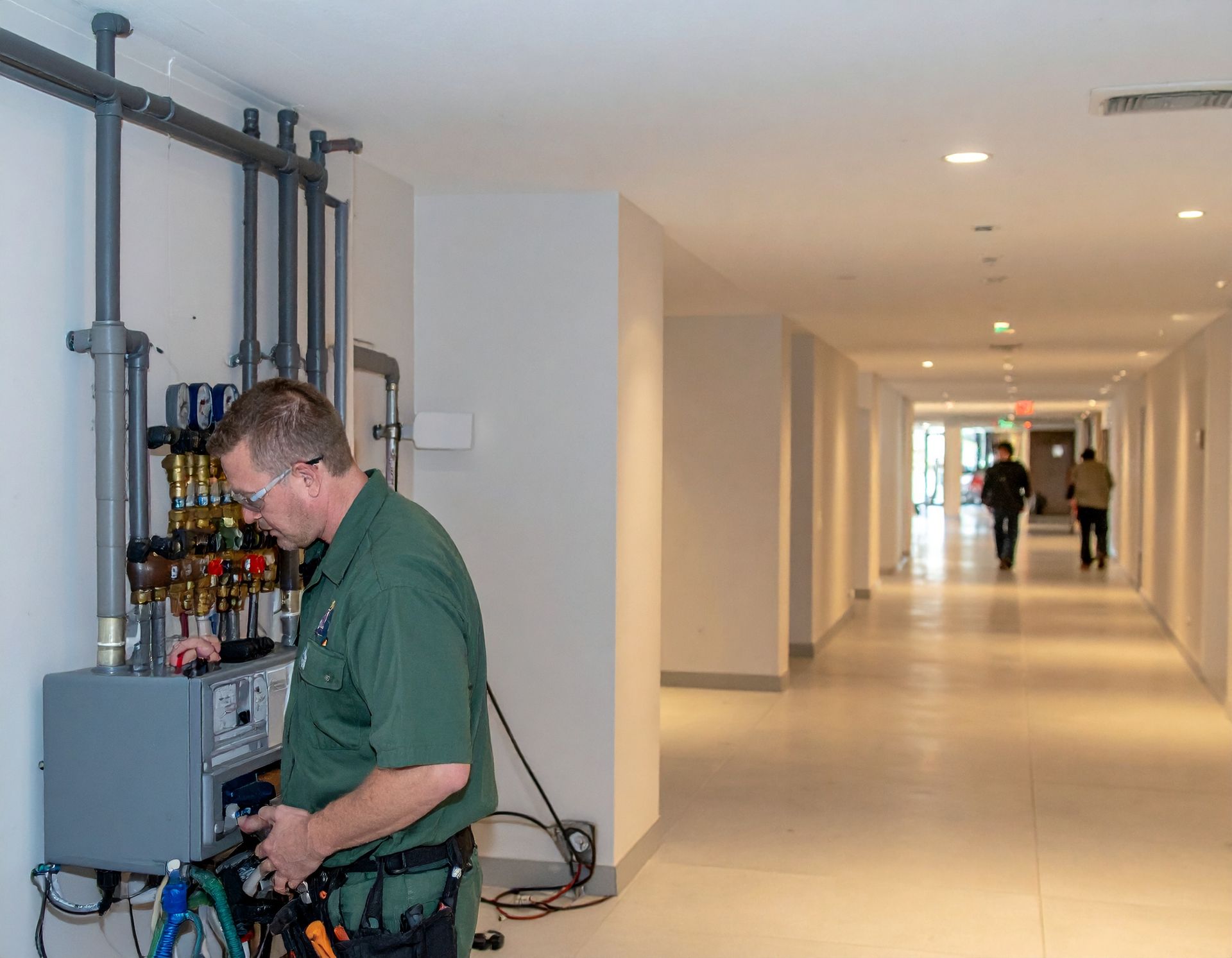 Technician adjusting cables beside equipment rack in a bright hallway; people visible in the distance.