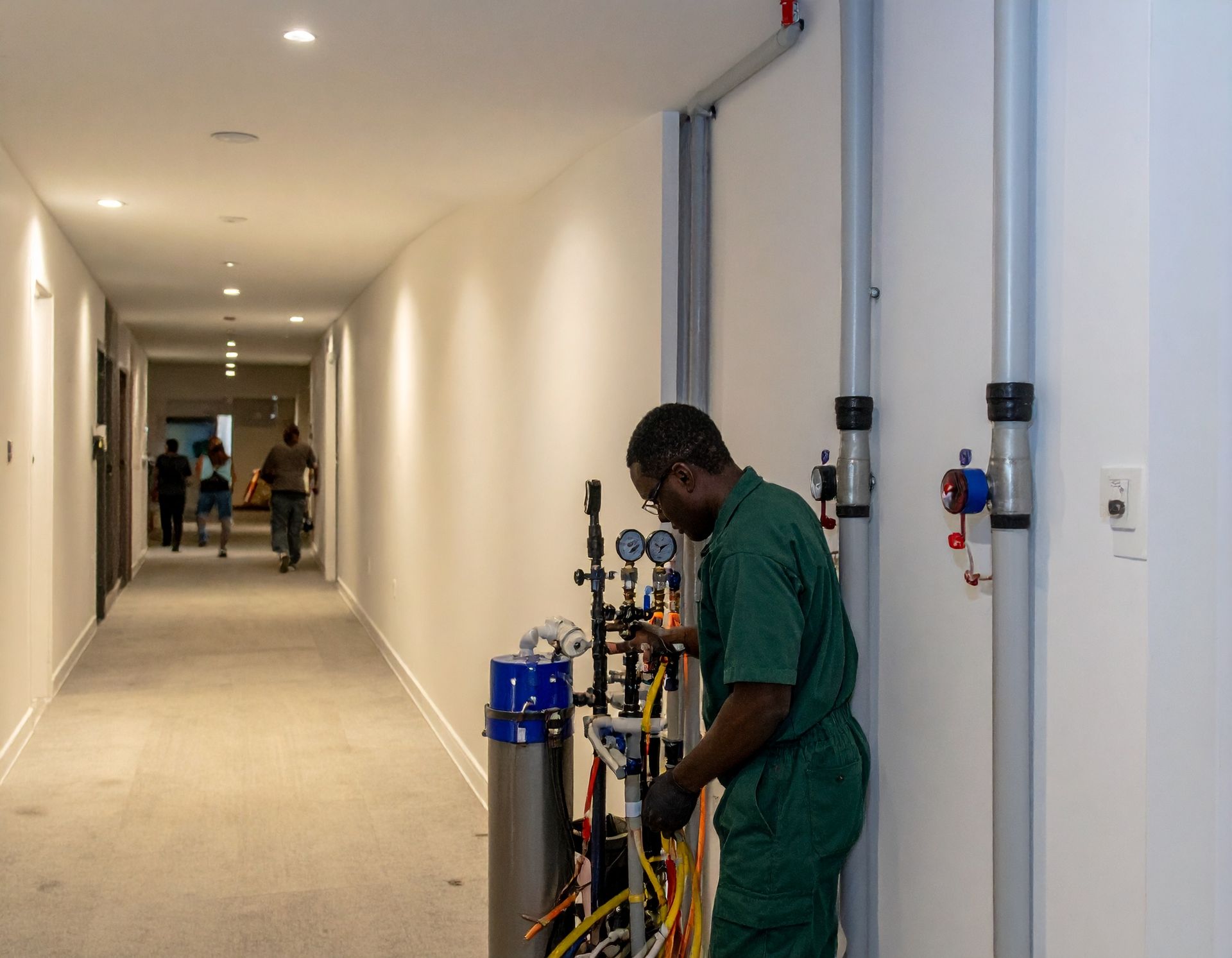Worker in green scrubs adjusting equipment in a long hallway with people in the distance.