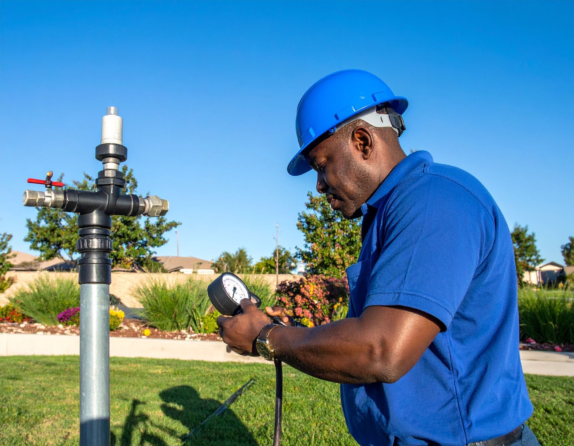Worker in blue hard hat inspecting a sprinkler valve in a landscaped yard.