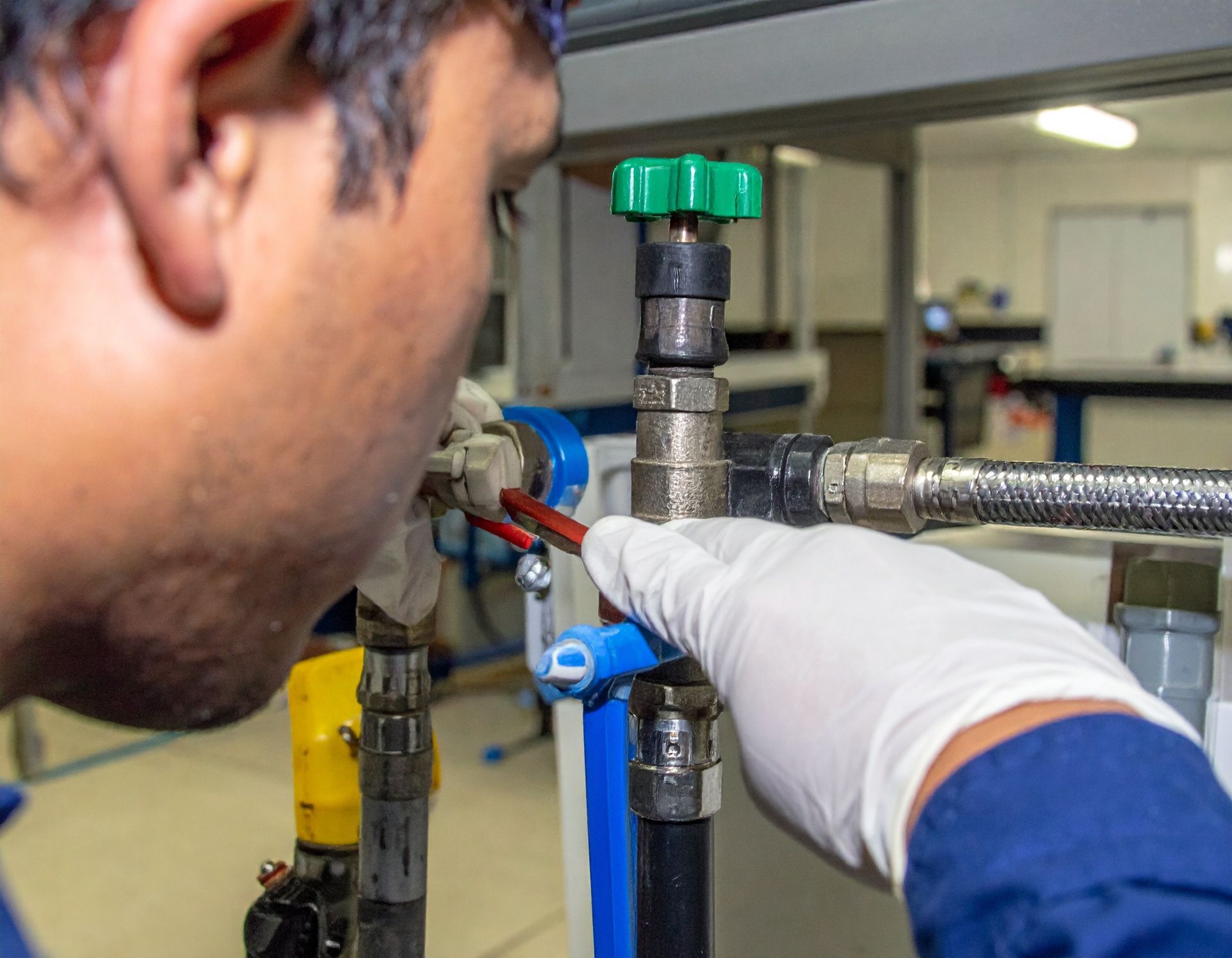 Gloved hand adjusting a valve on metal piping while a person watches in a workshop lab environment