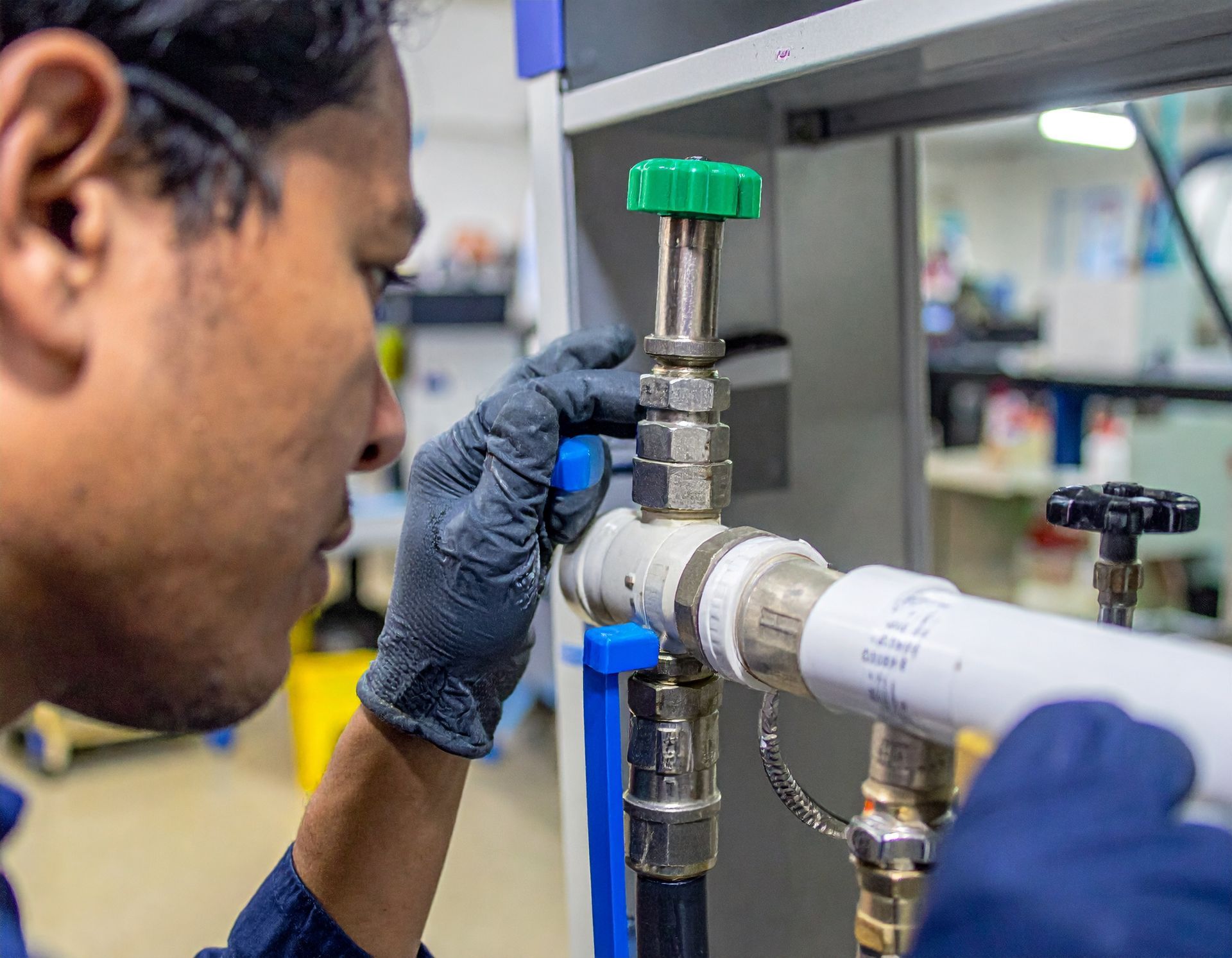 Technician adjusting a metal pipe assembly with a green valve in a lab or workshop.
