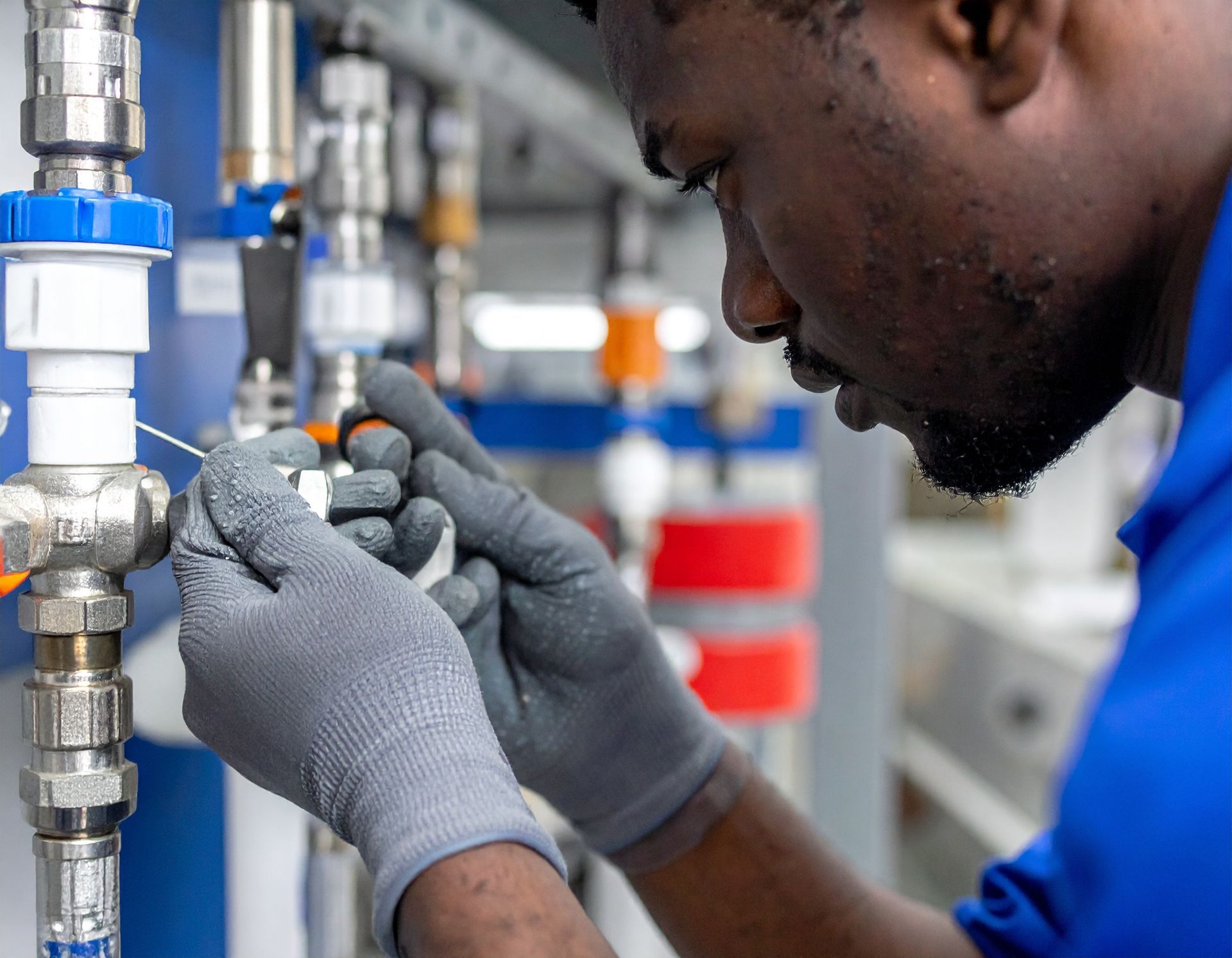Technician in blue uniform adjusts a valve on industrial piping with gloved hands.