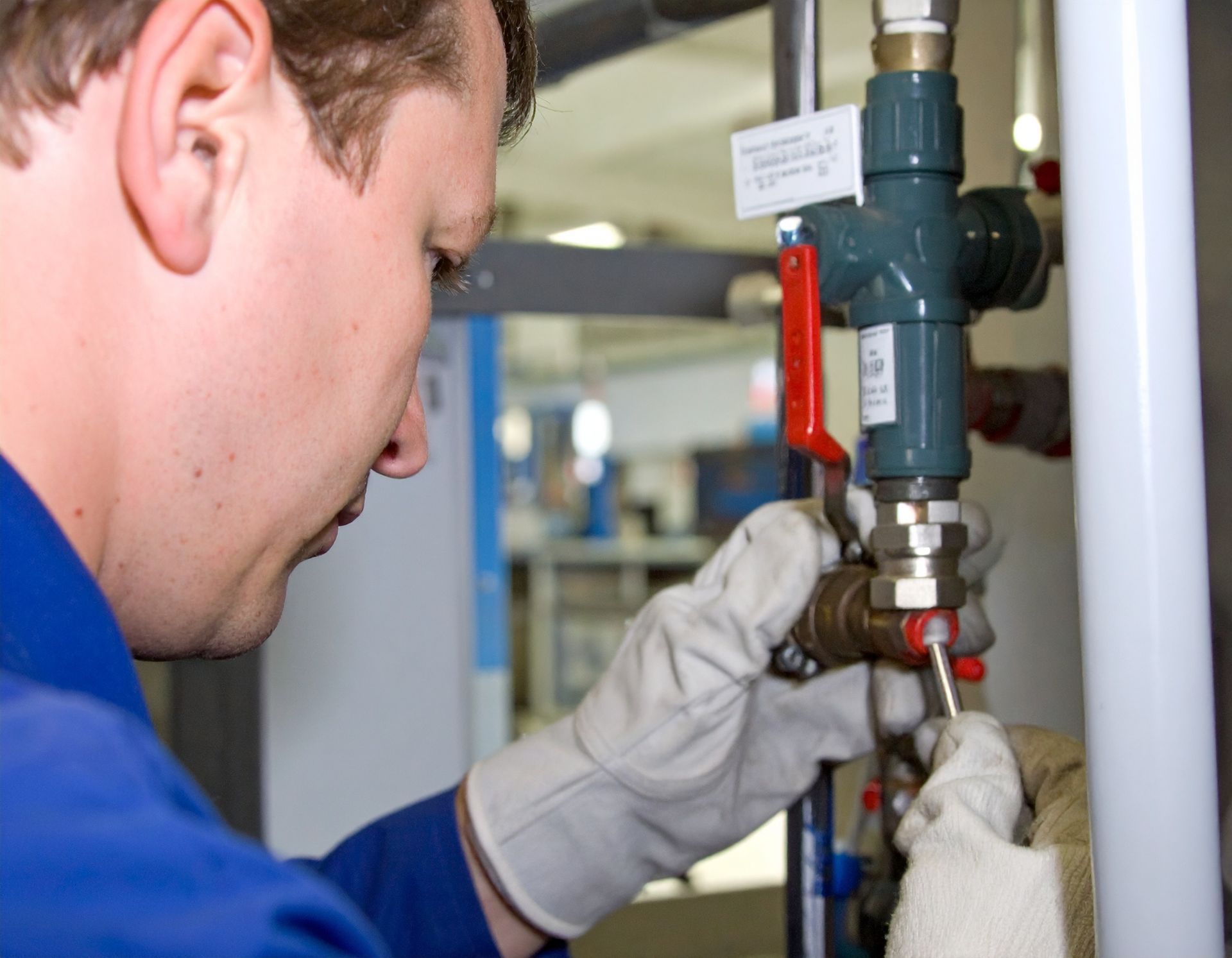 Technician in blue coveralls adjusts a valve on industrial piping, wearing gloves.
