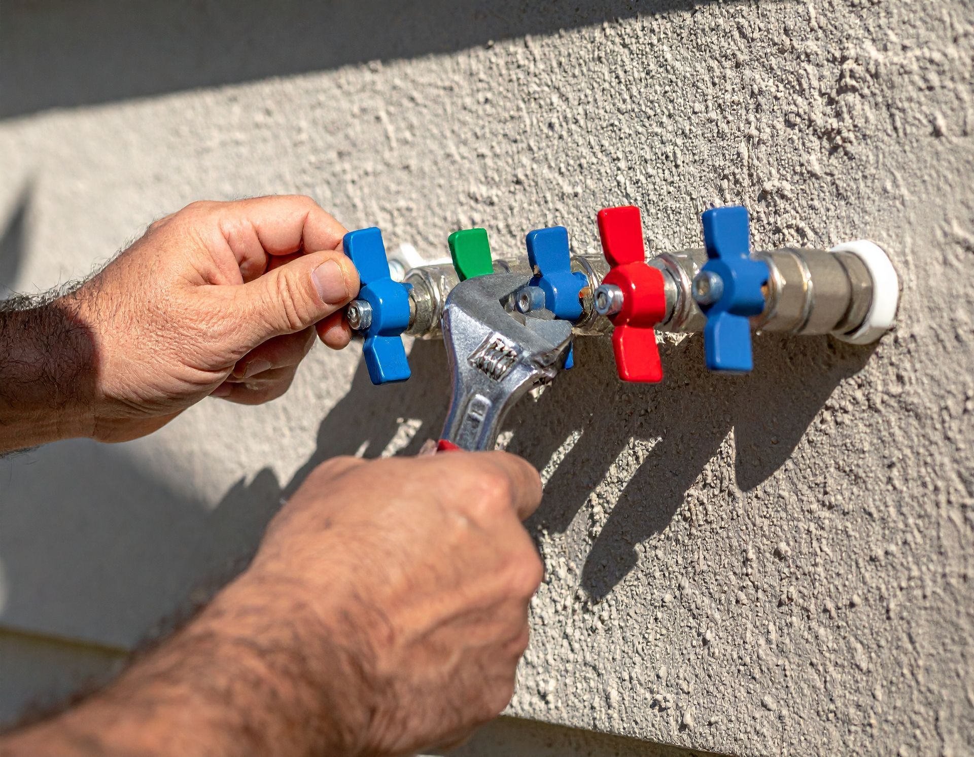 Hands using a wrench to tighten colorful hose bib handles on a wall-mounted outdoor water line