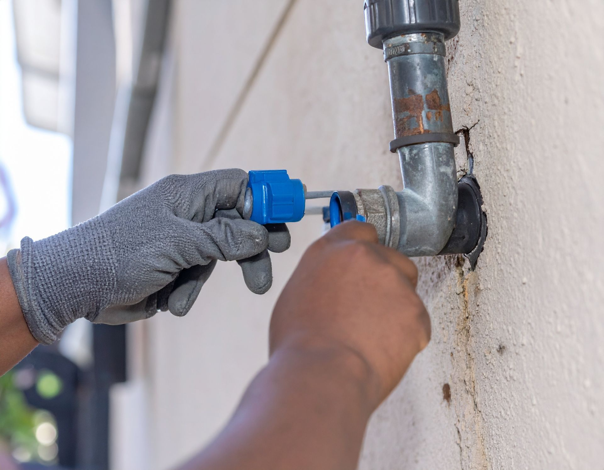 Hands using a blue valve wrench to tighten an outdoor water pipe connection on a wall.
