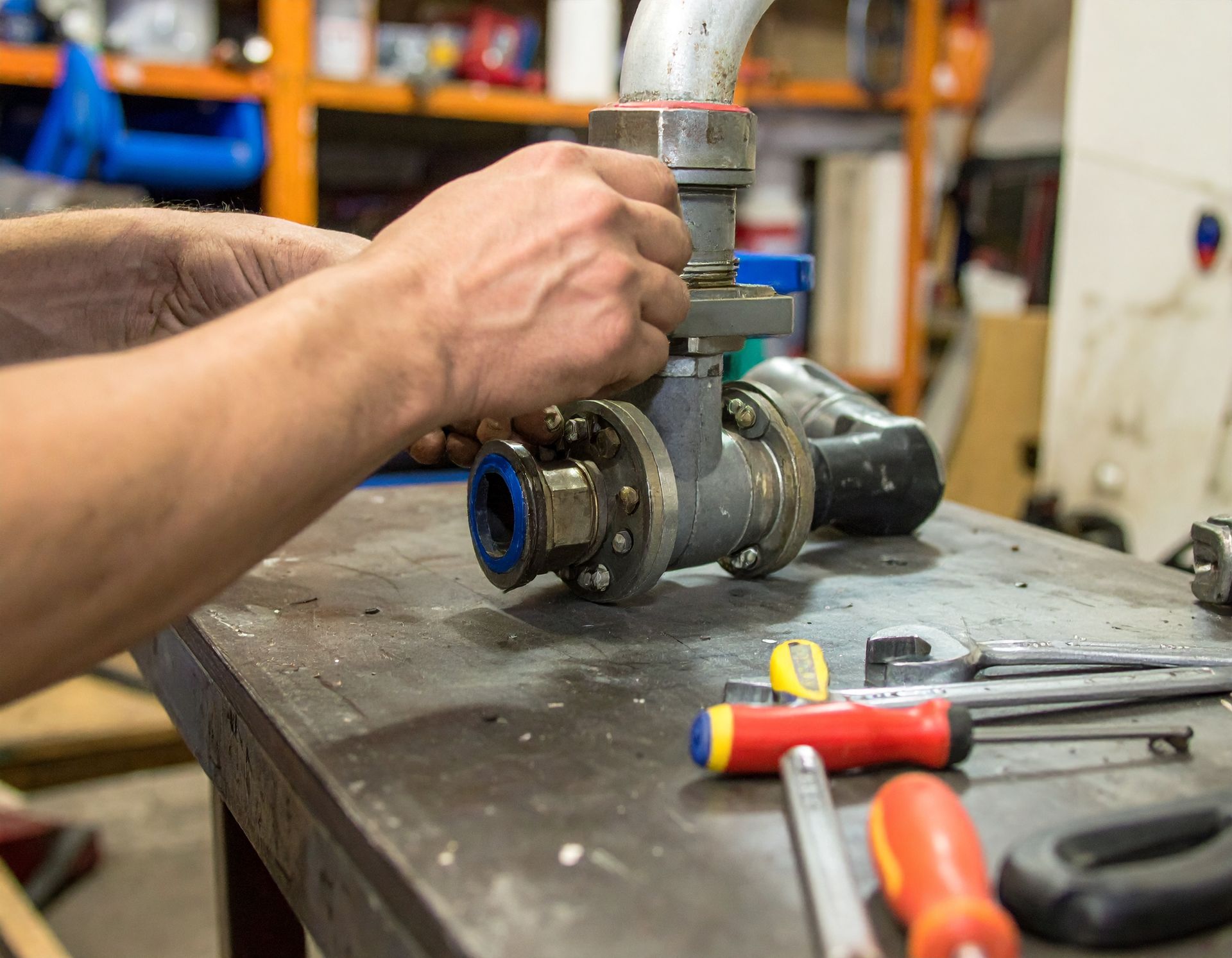 Hands repairing a metal pipe valve on a workbench with tools nearby.