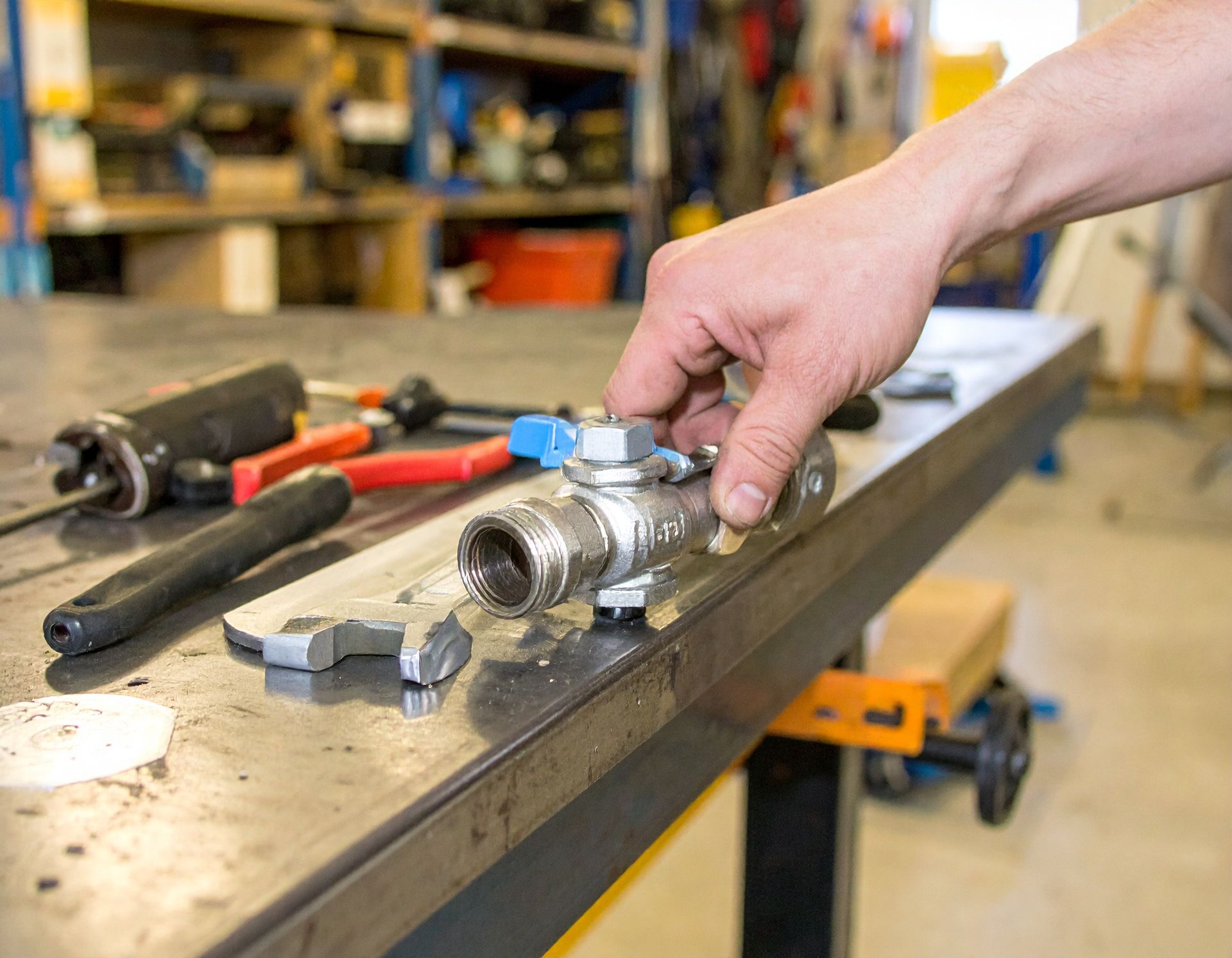Hand holding a metal tool part on a workbench in a workshop, with tools scattered nearby