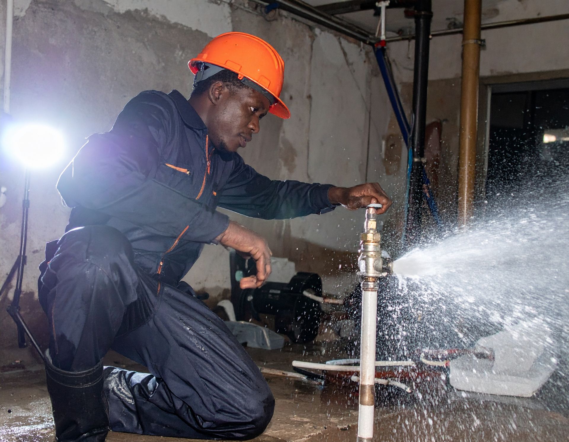 Worker in orange hard hat opening a pipe valve with water spraying in a dim industrial room