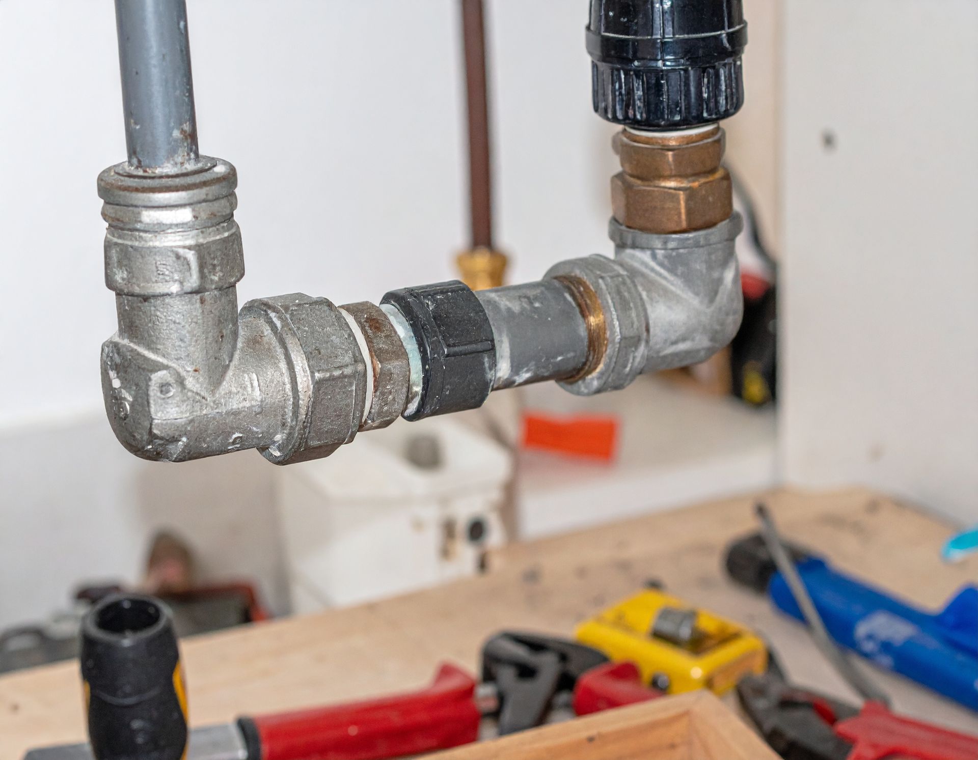 Metal plumbing pipes and fittings under a sink, with tools on the floor in the background