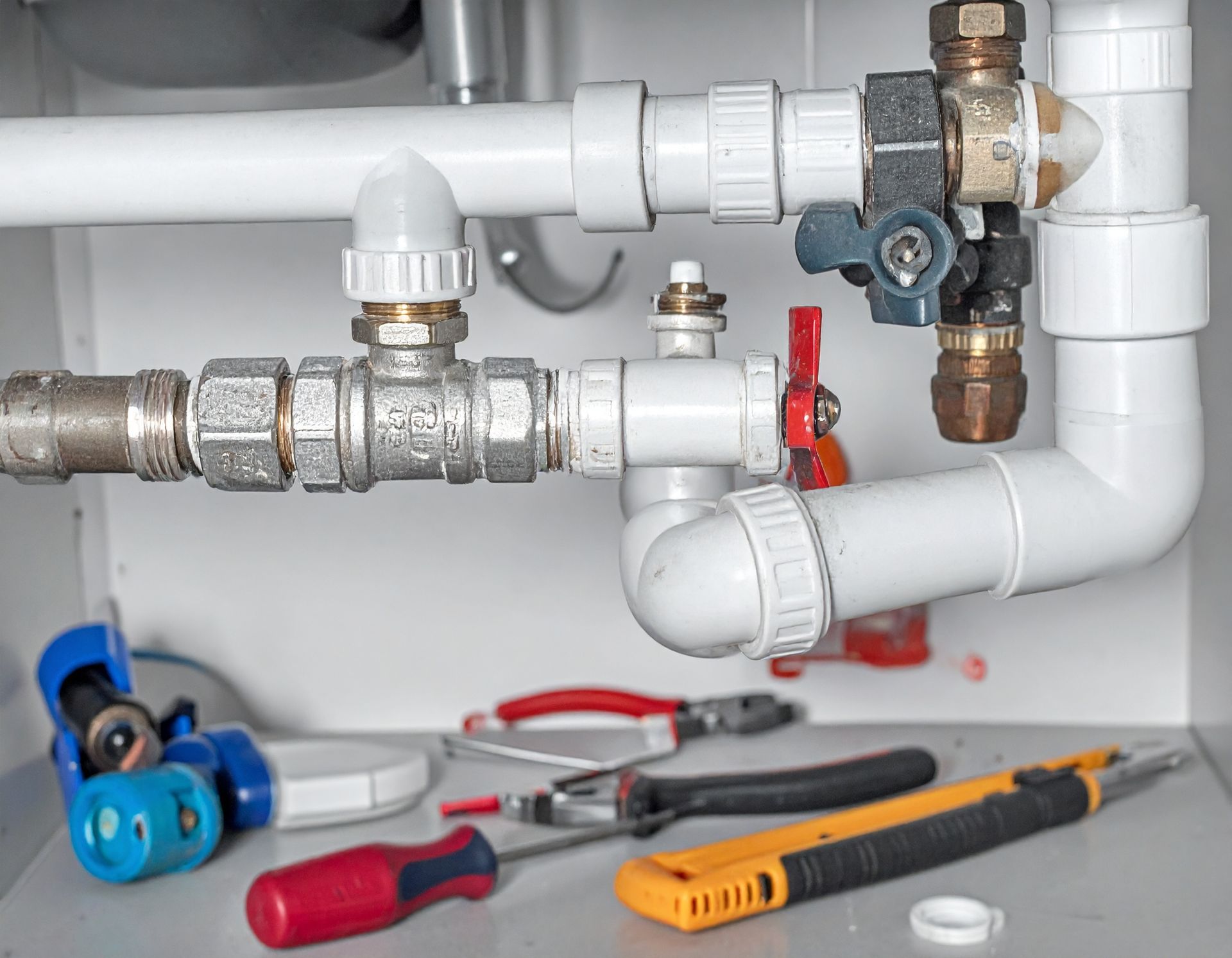 Under-sink plumbing with pipes, valves, and hand tools laid out for repair on a white floor