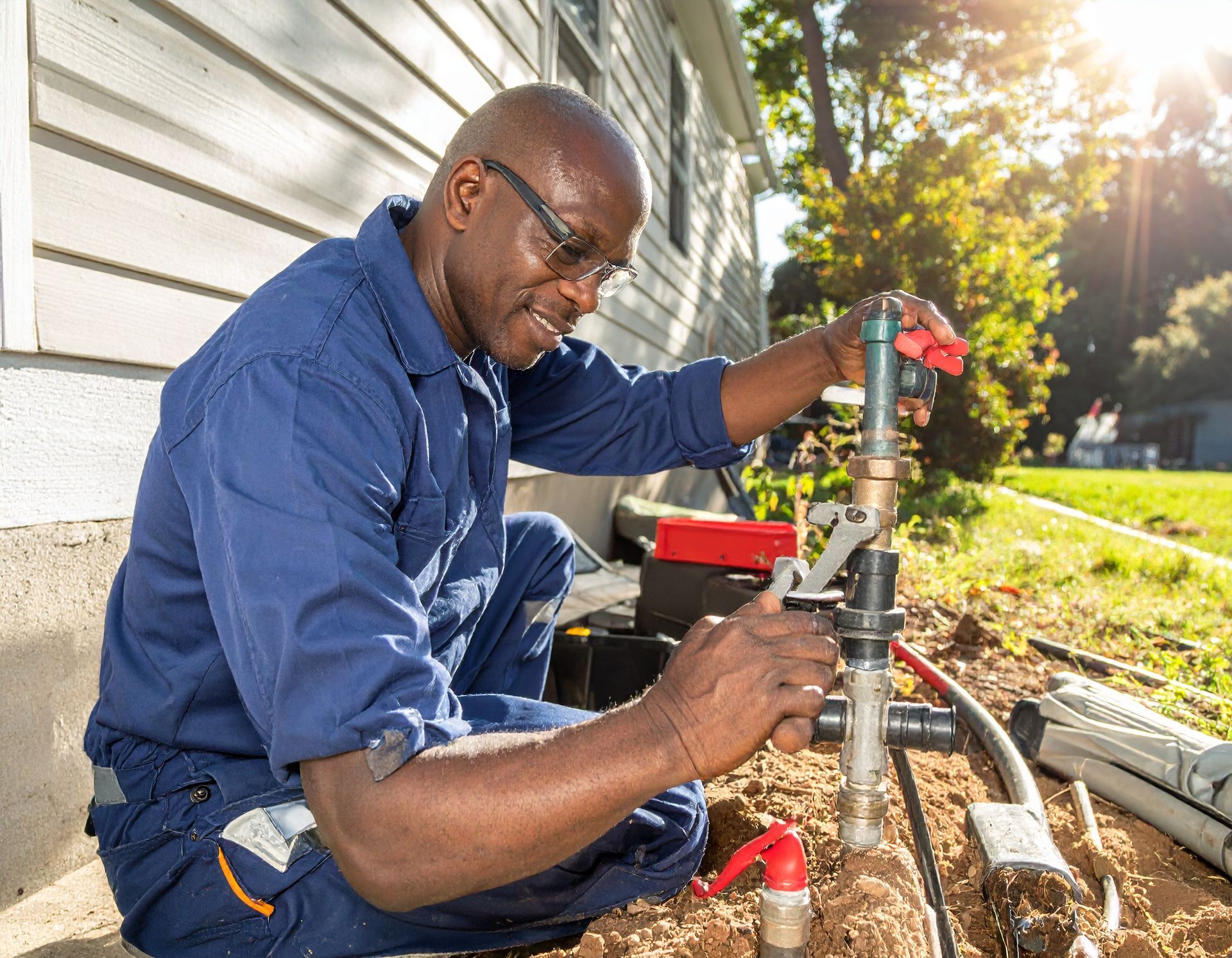 Worker using a pipe wrench to fix outdoor plumbing beside a house in bright sunlight