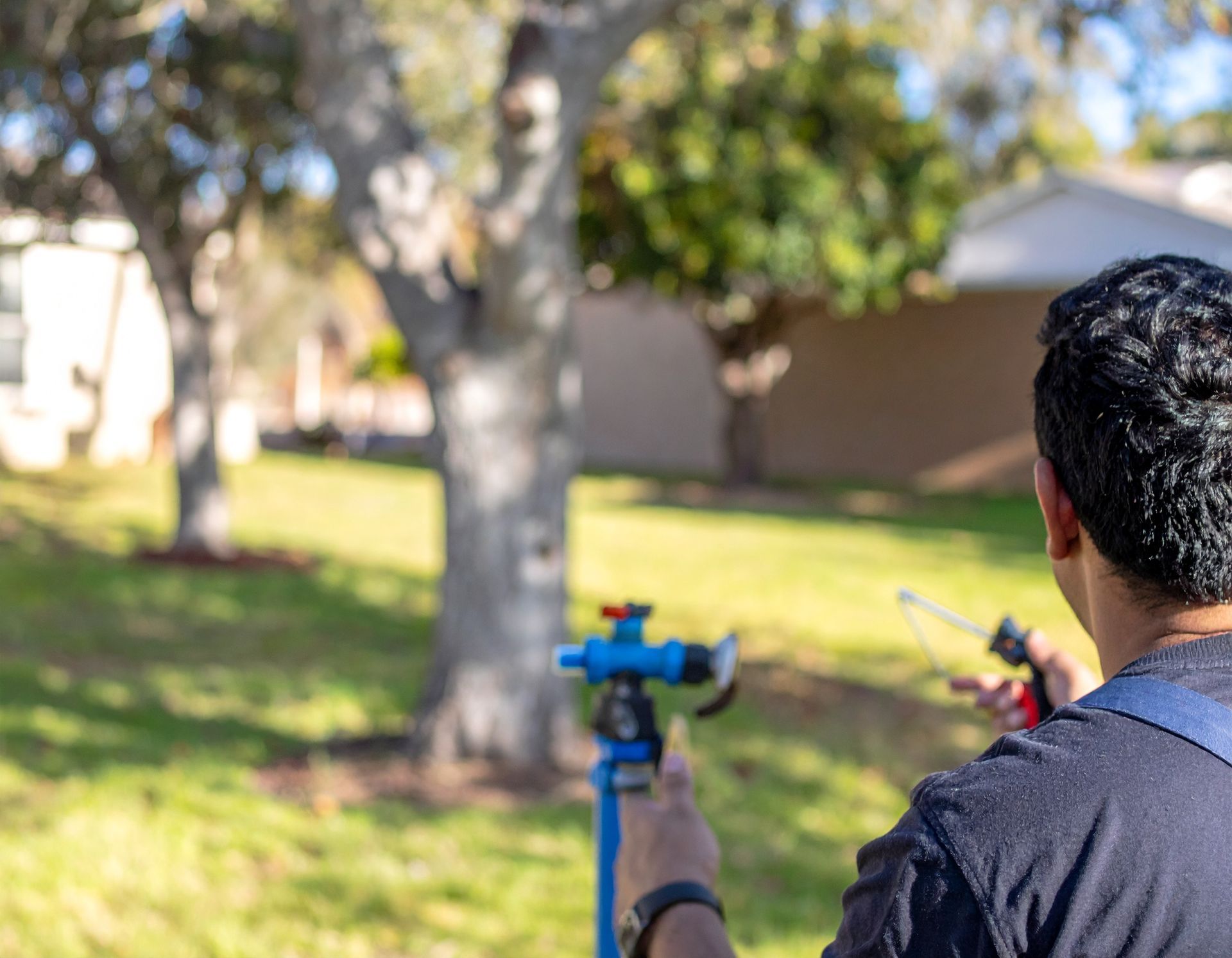 Person using a blue spray gun in a sunlit backyard near trees and a house