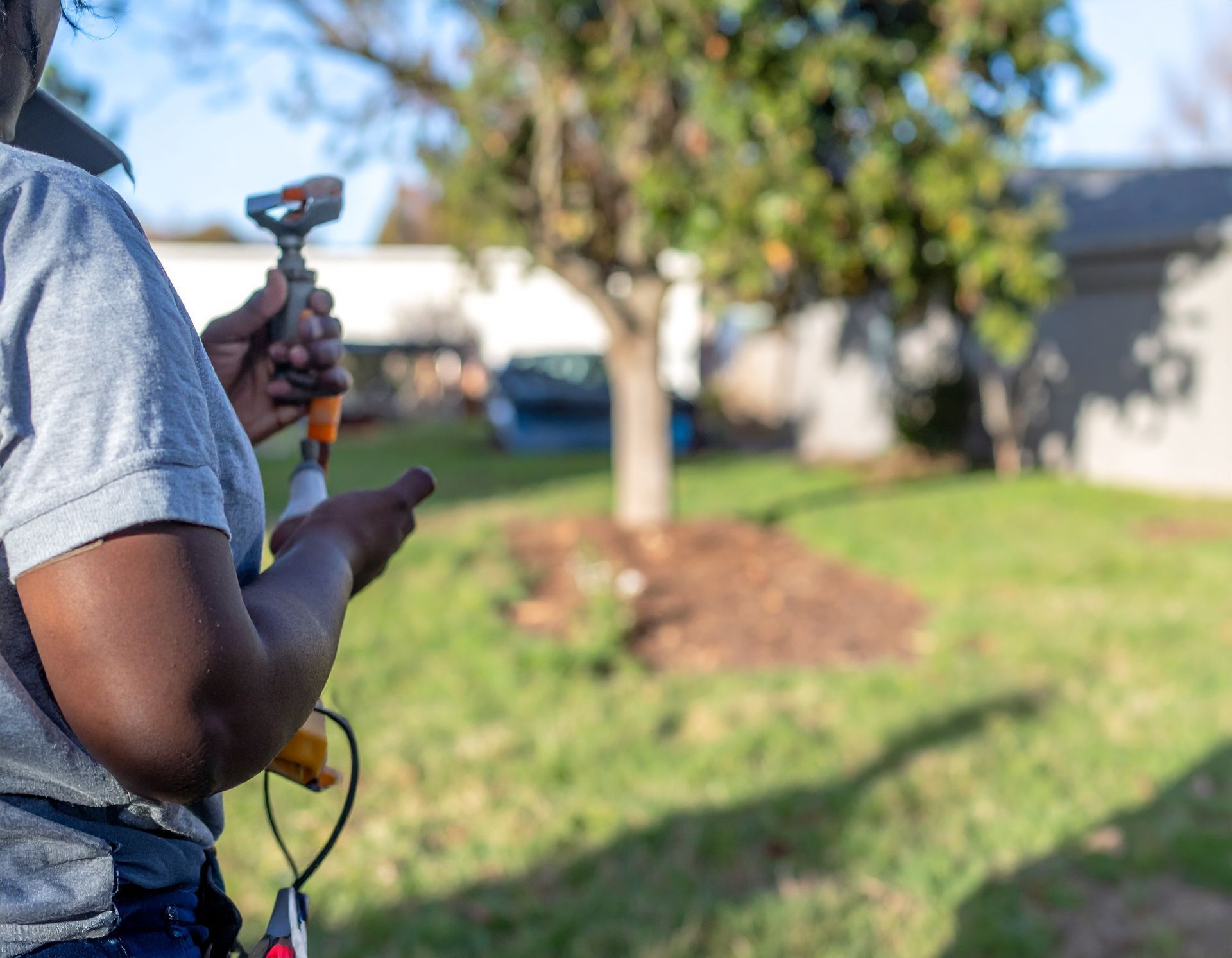 Person holding a camera on a tripod in a sunny yard with a tree and house in the background