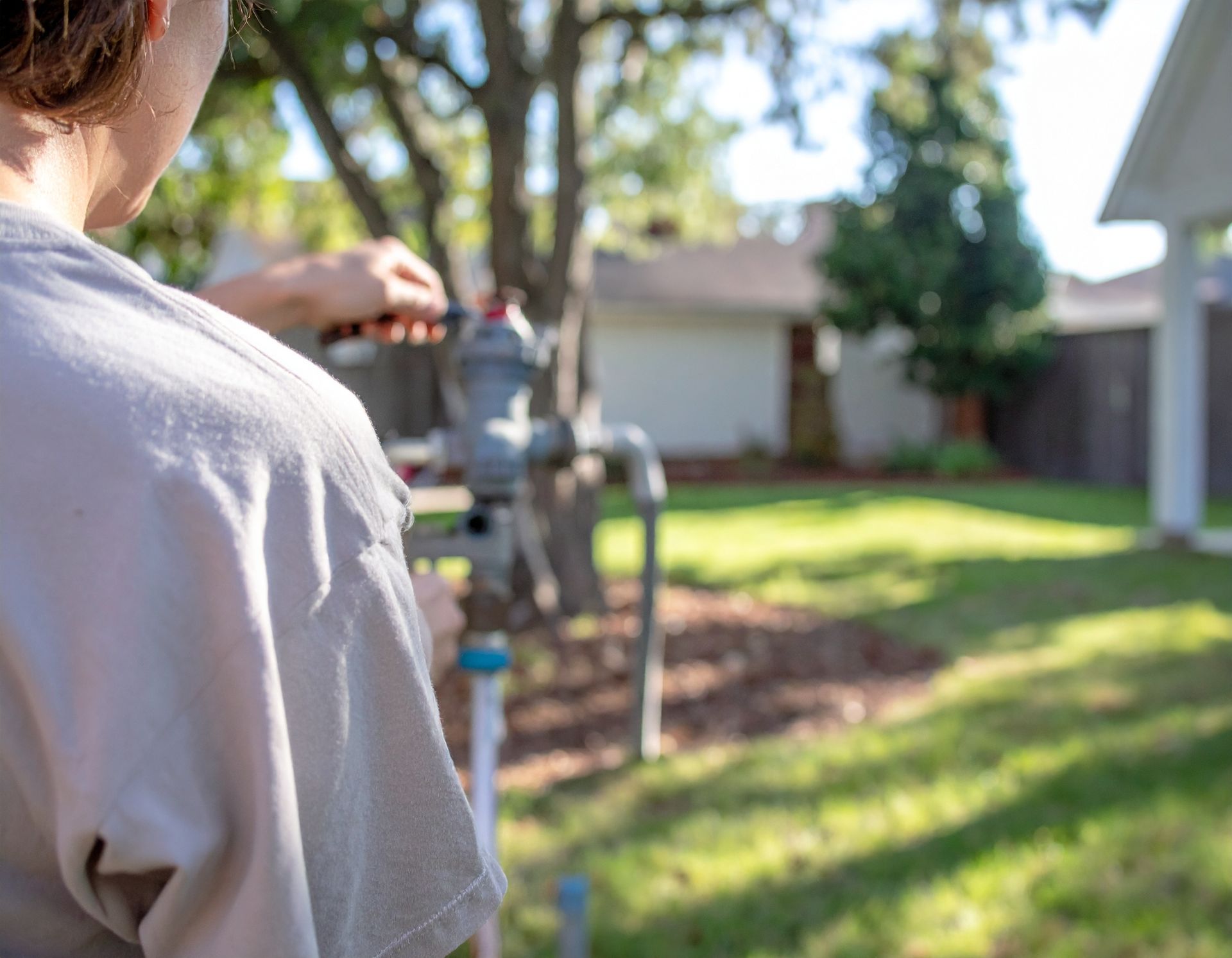 Person in a light shirt aiming a garden hose in a sunny backyard.