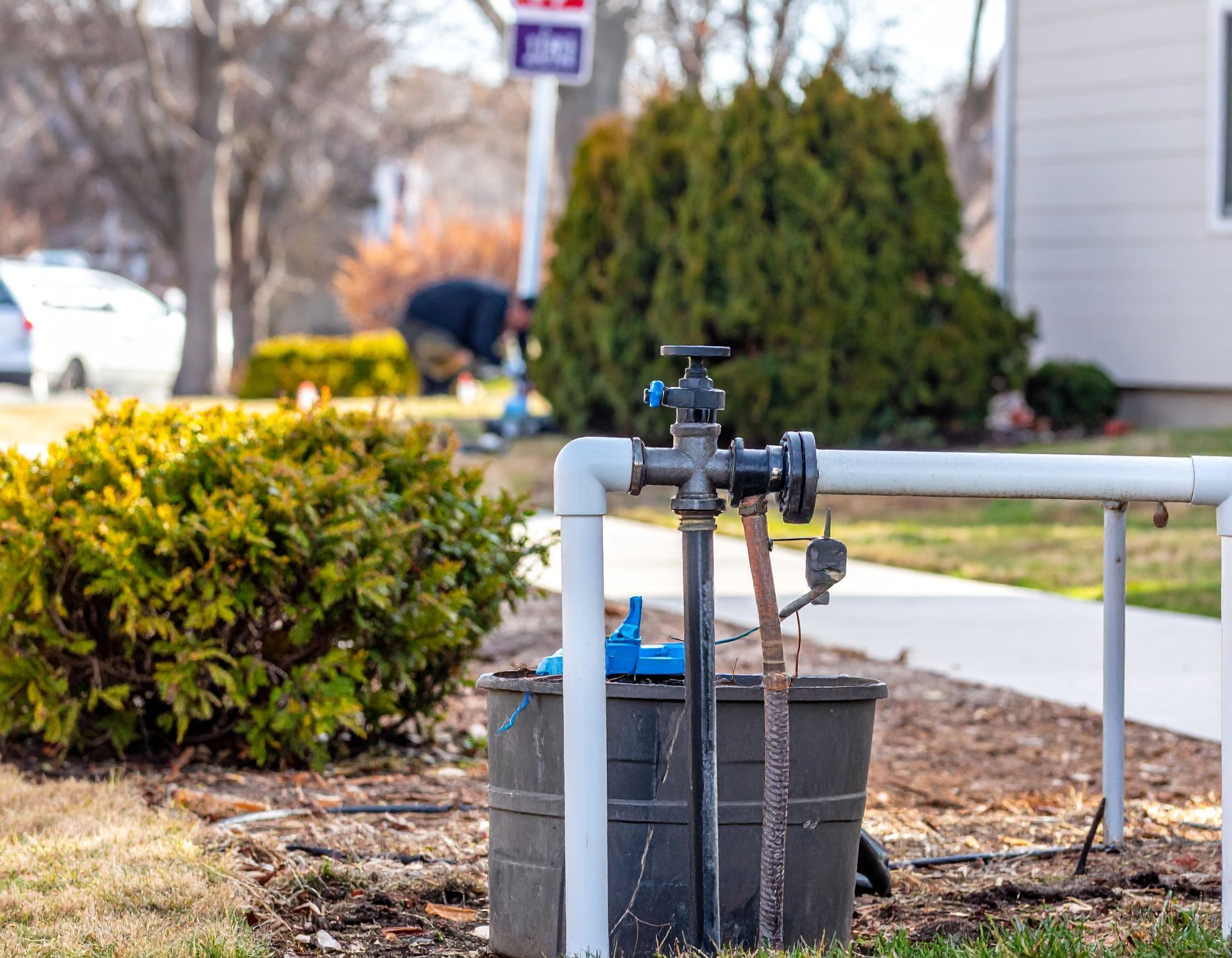 Outdoor water spigot and hose valve beside a sidewalk, with shrubs and a house in the background