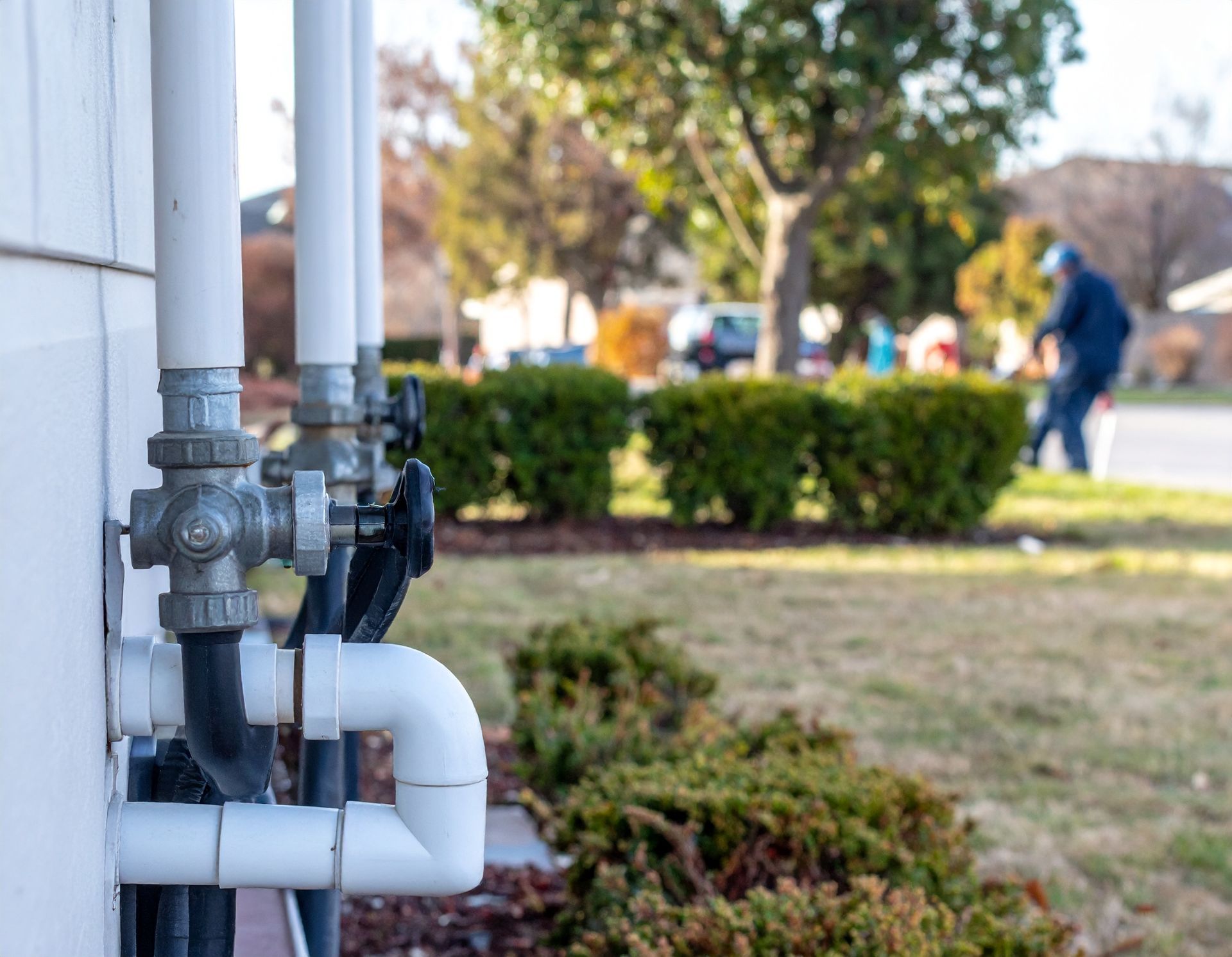 White PVC pipes and a gas meter mounted on a house exterior beside a lawn and shrubs.