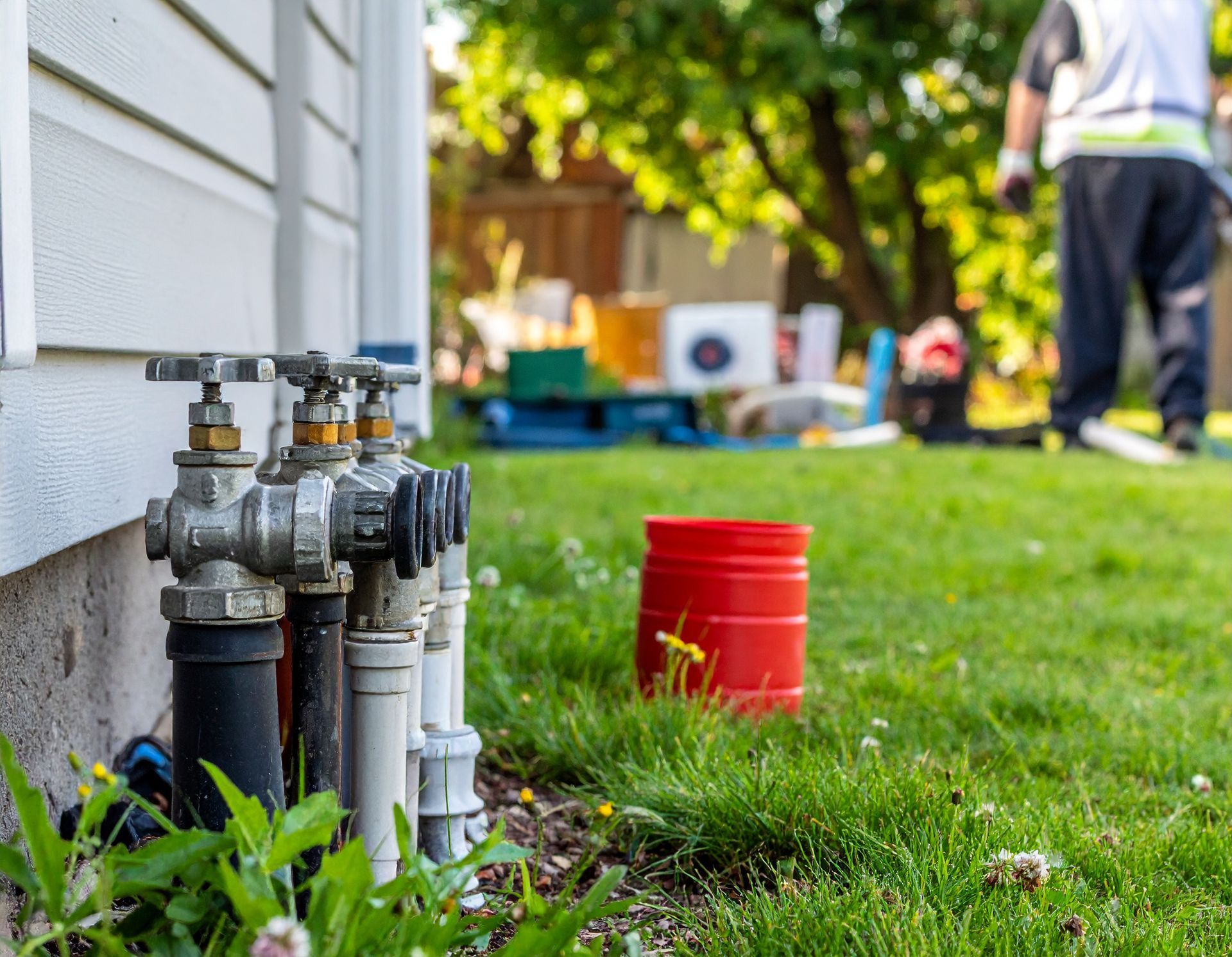 Outdoor water valve assembly beside a house, with a red bucket and worker in the background on a grassy lawn