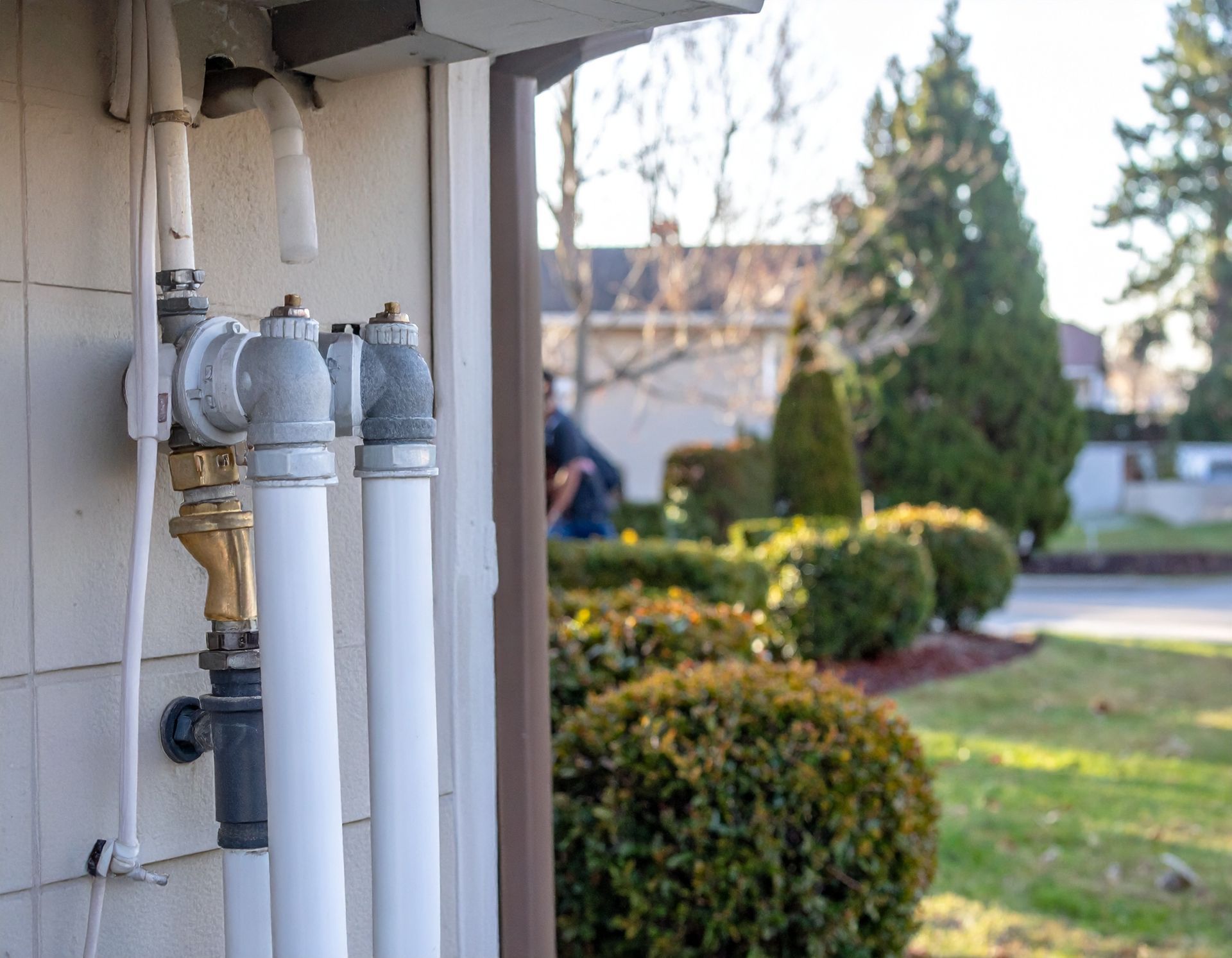 Utility meters and white pipes beside a house, with a sunlit yard and shrubs in the background
