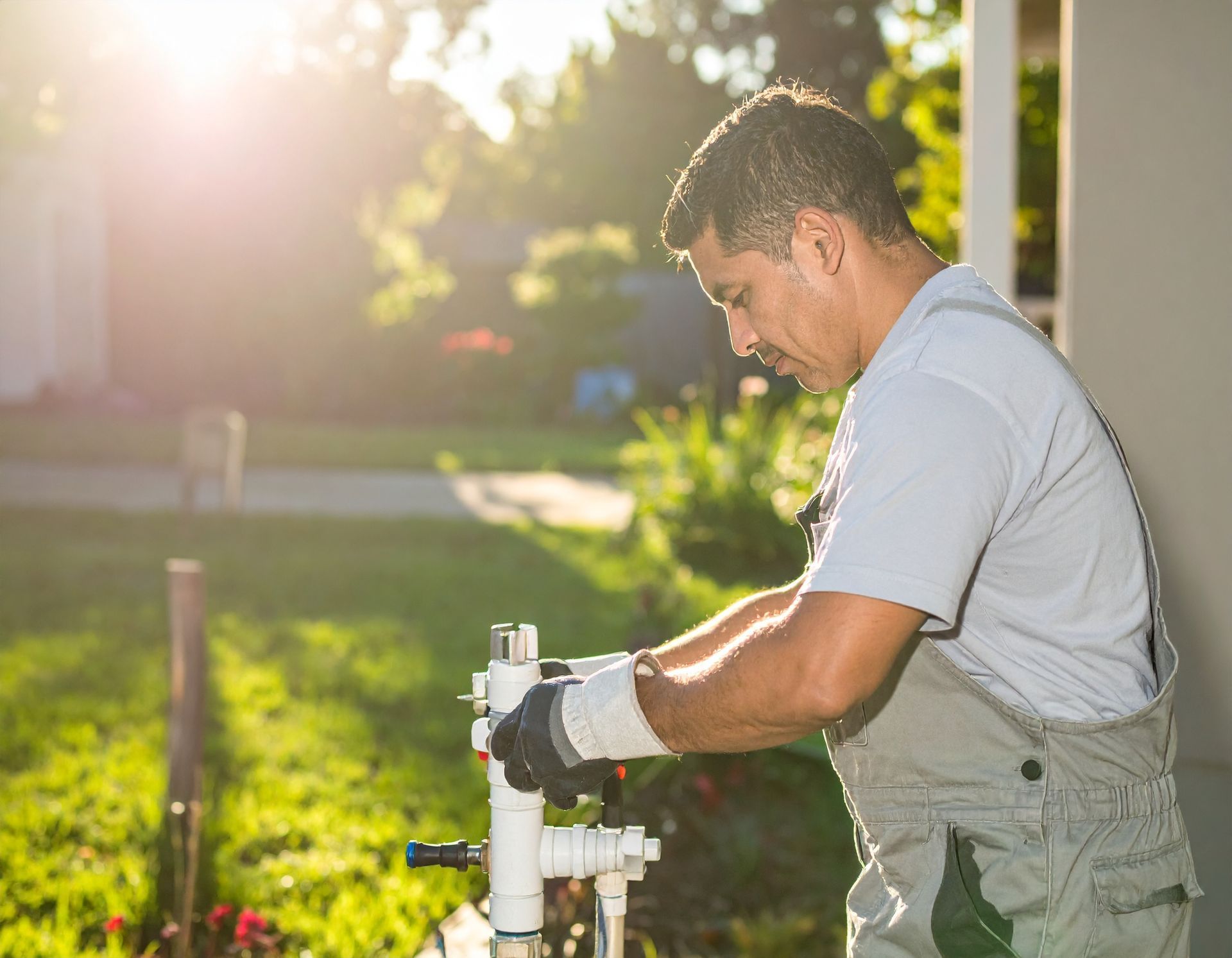 Worker in gray overalls fixing a white pipe outdoors in bright sunlight.