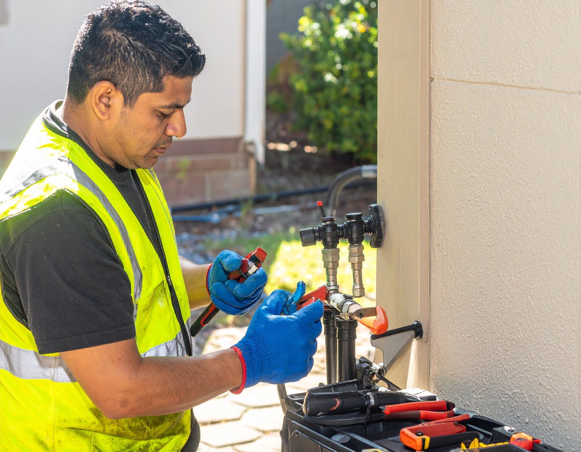 Worker in a neon safety vest using pliers on outdoor plumbing equipment beside a building wall