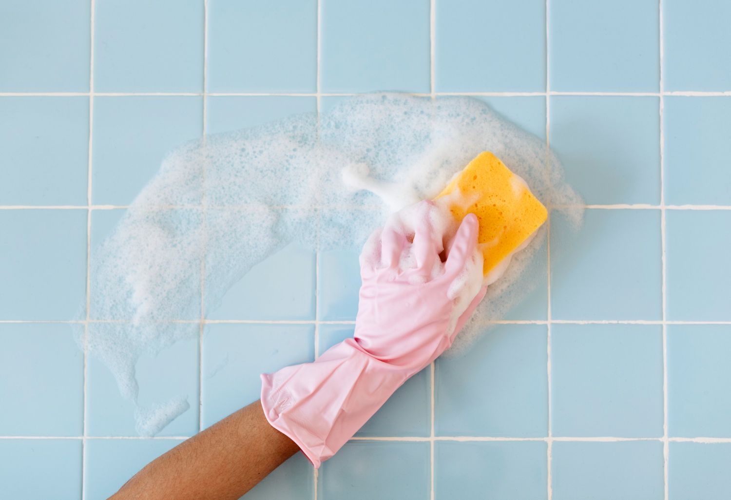 A person wearing a pink glove is cleaning a tiled wall with a sponge.
