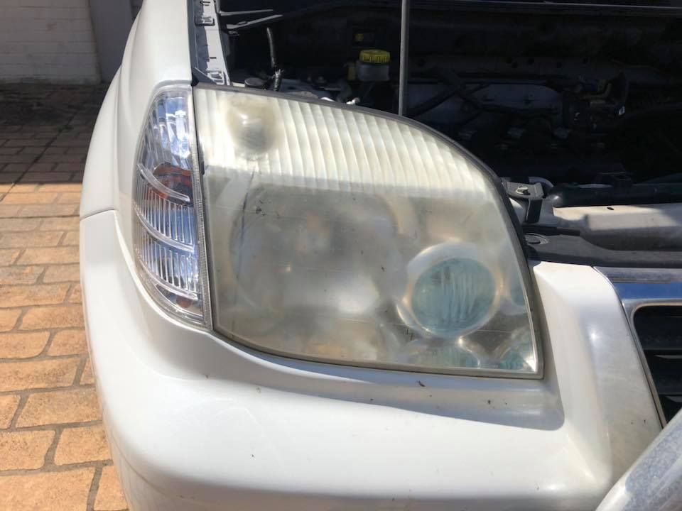 Close-up of a white car’s front left headlight and grille with the hood open.