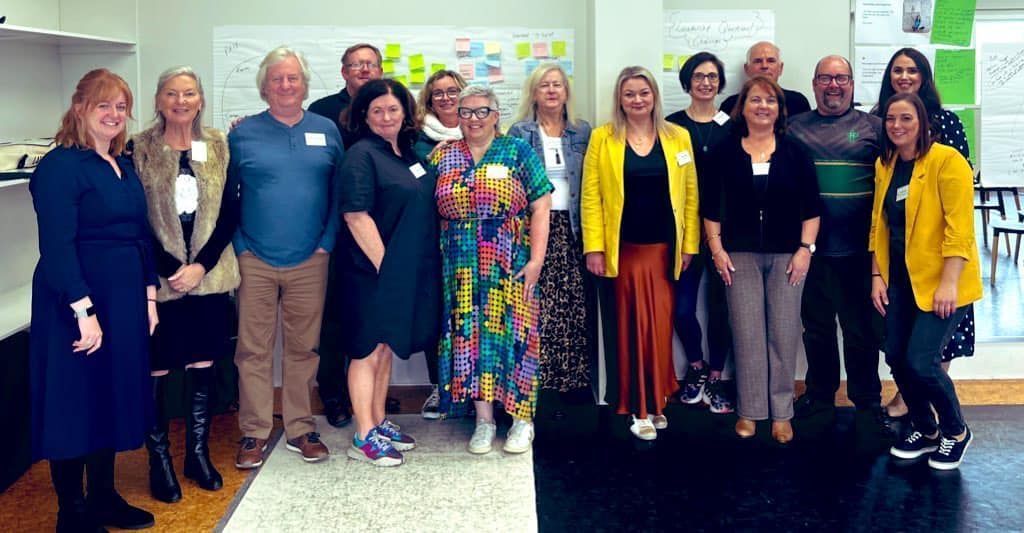 A diverse group of fourteen people stand in two rows smiling for a photo in an office setting with wall notes in the back.