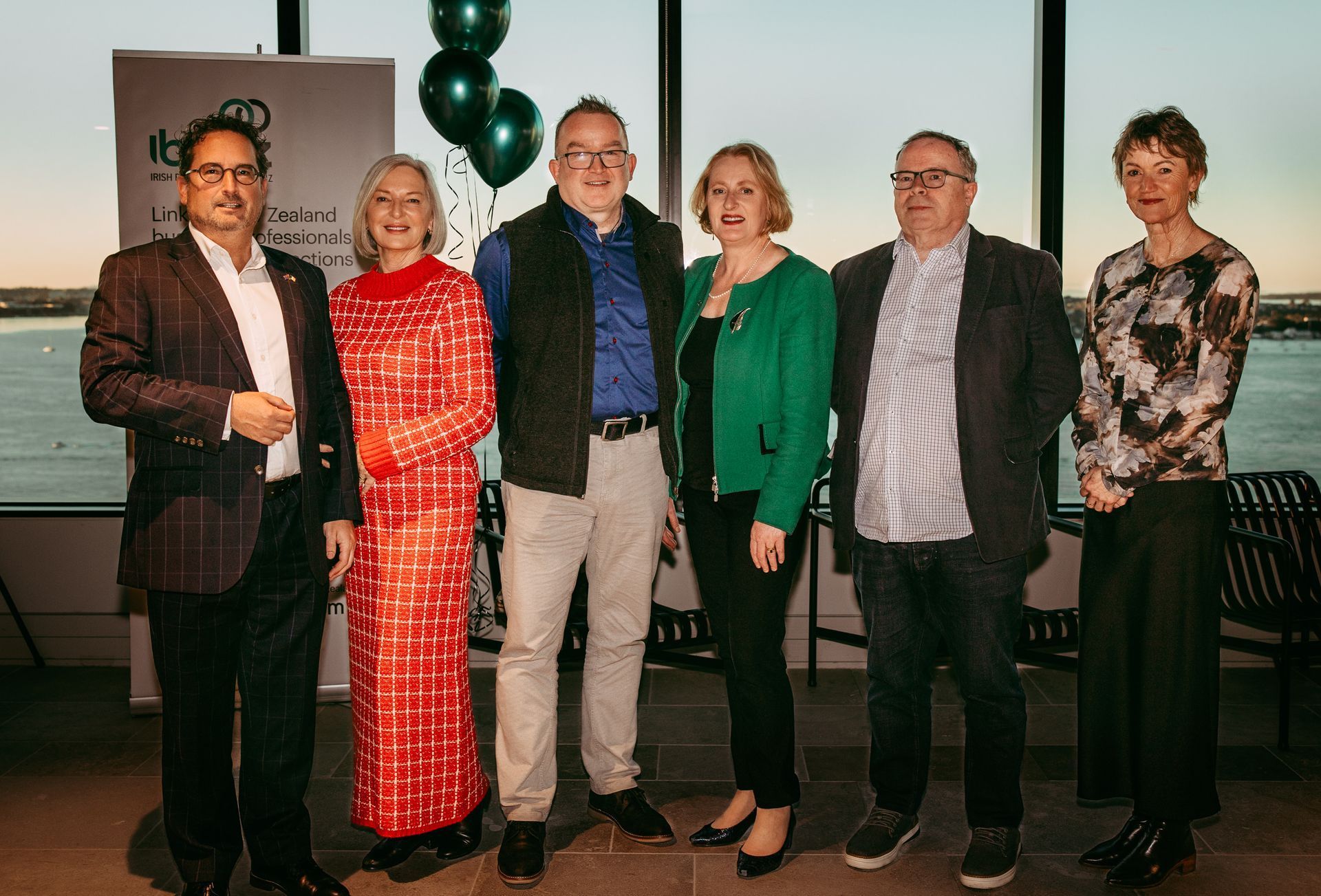Six people stand in a line posing for a photo indoors, with a water view and green balloons in the background.