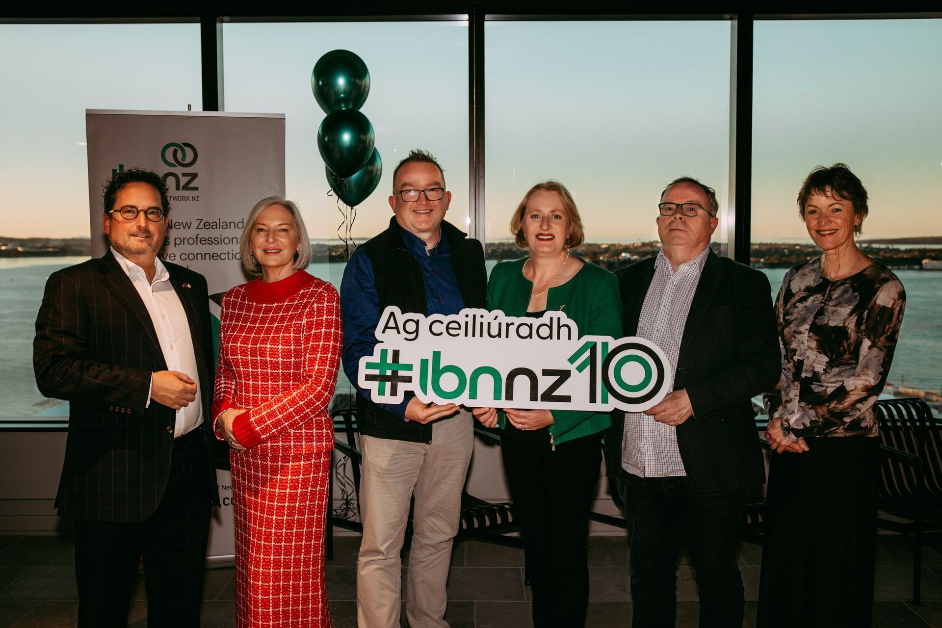 Six people stand in a line against a window at sunset, holding a sign that says #ibnnz10, with green balloons in the back.
