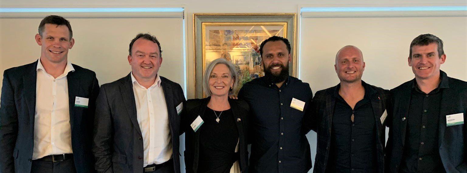 Six people stand in a line against a light-colored wall with a framed picture between them, all wearing business attire.