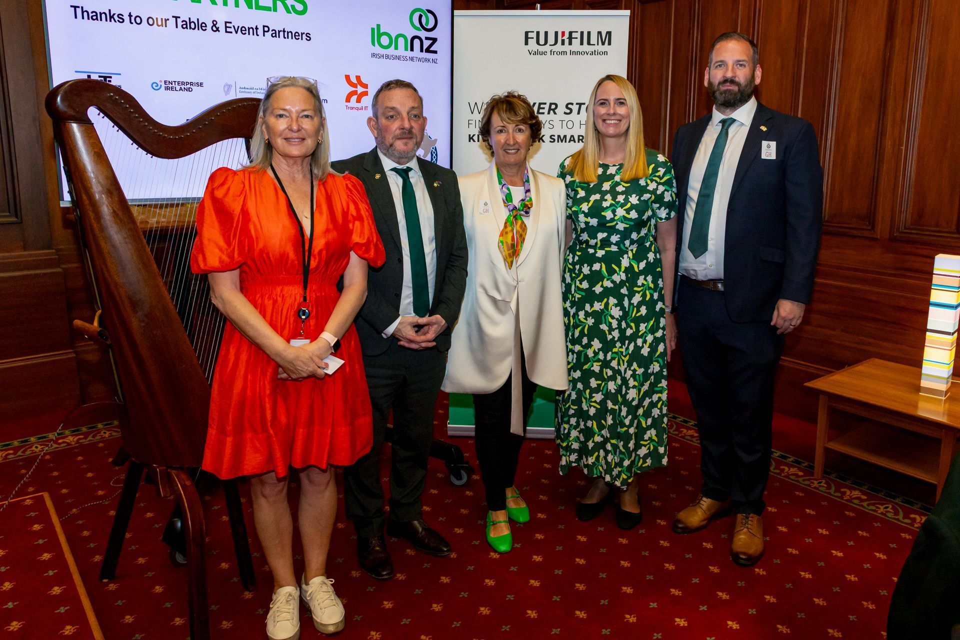 Five people stand together in a row indoors in front of a harp and a sponsor banner, smiling for a professional photo.