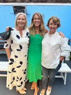 Three people pose together outdoors against a blue wall, smiling while wearing casual outfits.