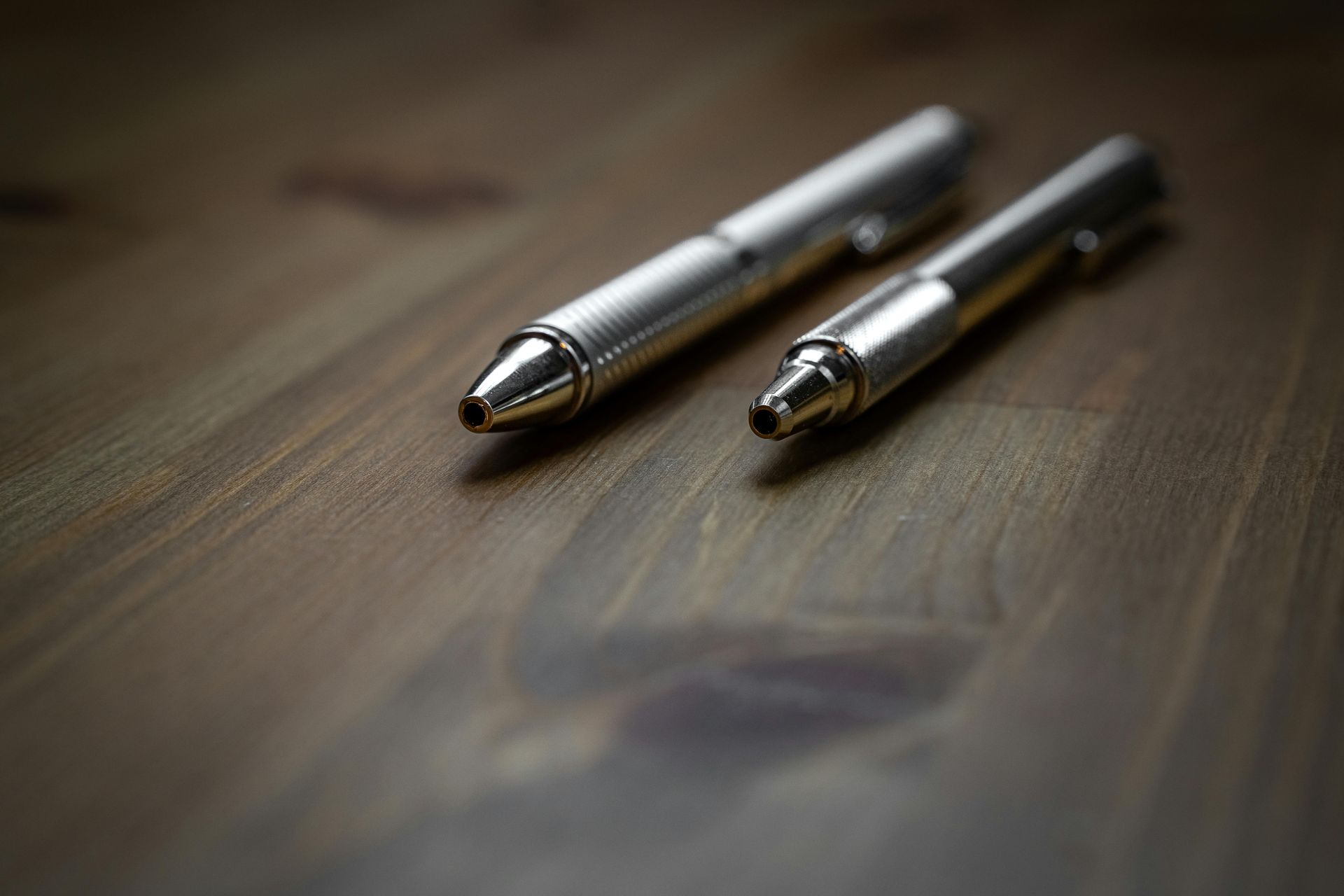 Two metallic pens resting on a dark, wood-grain surface.