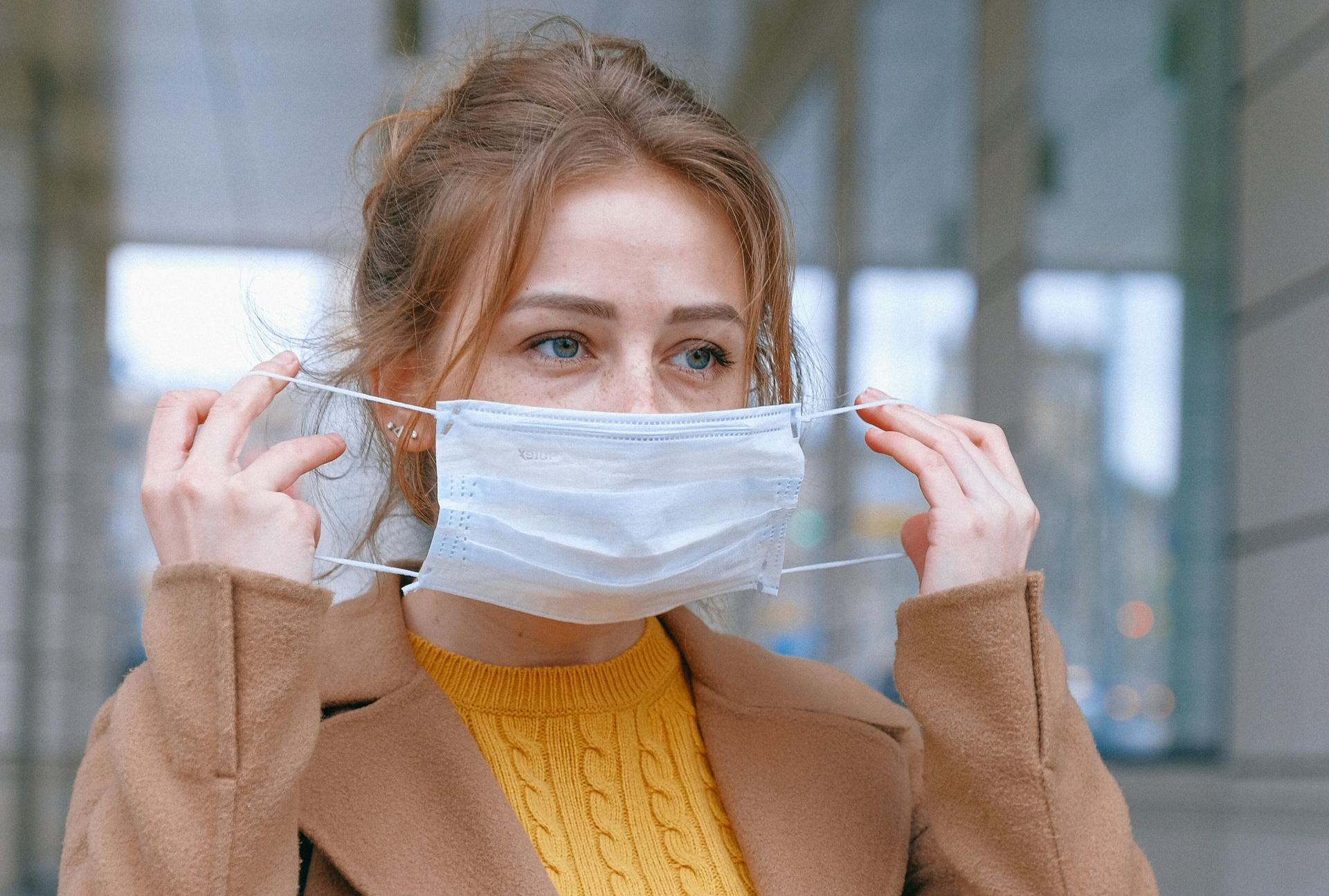 A person wearing a mustard yellow sweater and tan coat pulls a white face mask over their nose and mouth.