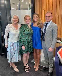 Four people stand smiling together in an indoor office setting, three wearing dresses and one in a suit and tie.