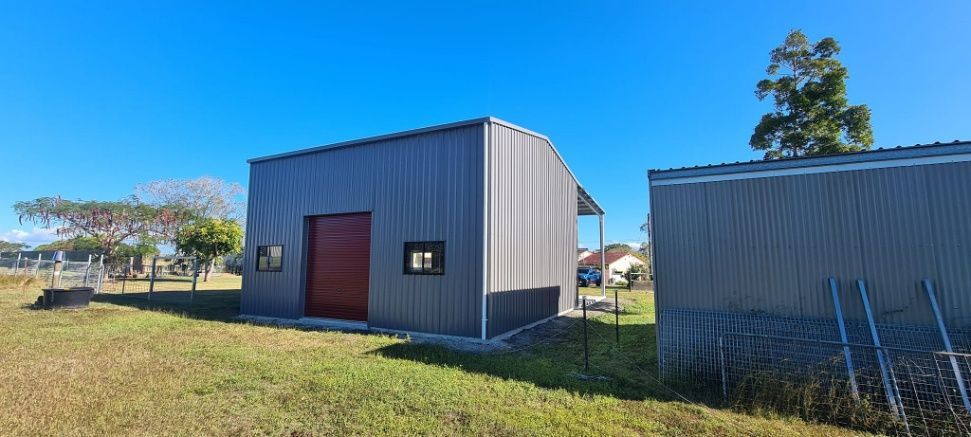 A Metal Building With A Red Door Is Sitting In The Middle Of A Grassy Field — Bushman Sheds in Bundaberg East, QLD
